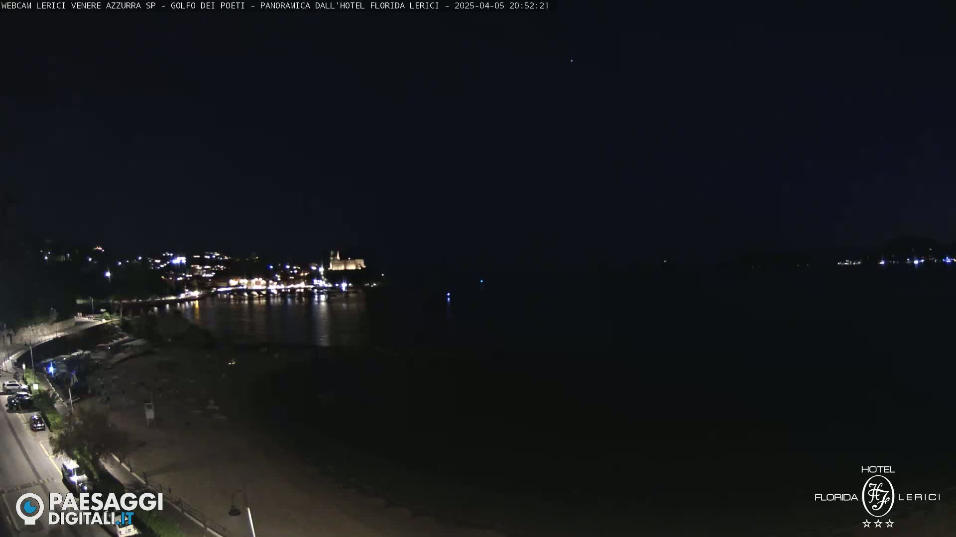 A nighttime coastal scene shows a dark, calm body of water reflecting the lights of a town across the bay, under a clear, dark sky.