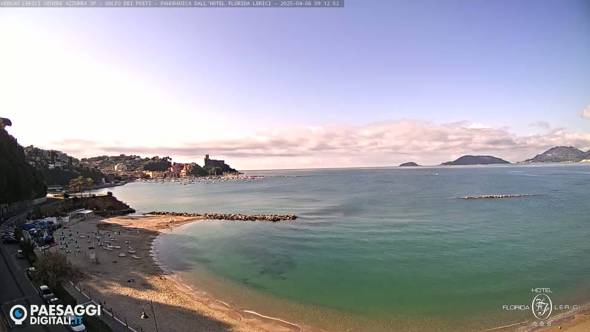 A partly cloudy day shows a sandy beach with several boats in the calm, teal water, a town on a hillside, and islands in the distance.
