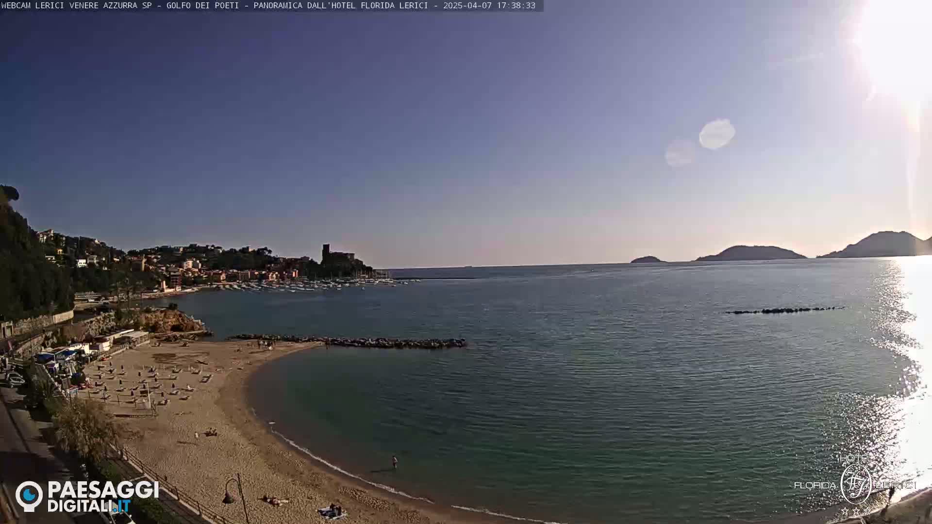 A sandy beach curves into a calm bay, overlooked by a town nestled against hills, under a clear, sunny sky.