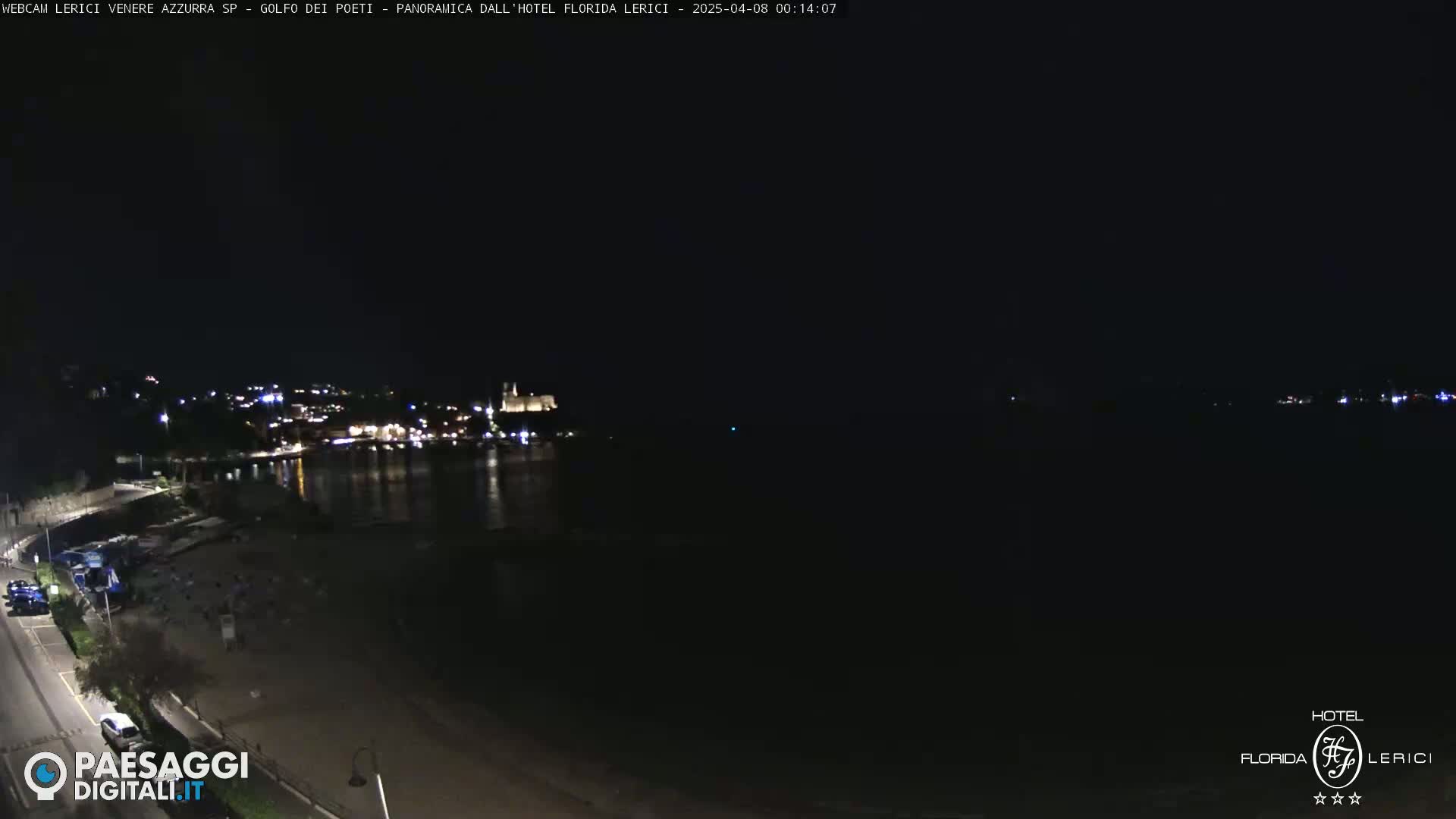 A nighttime view of a coastal town with lights illuminating the buildings and shoreline, under a dark sky.