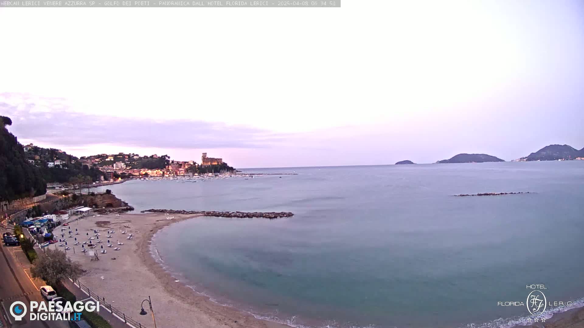 A sandy beach with lounge chairs sits beside a calm sea under a pale, twilight sky, with a town and castle visible in the distance.
