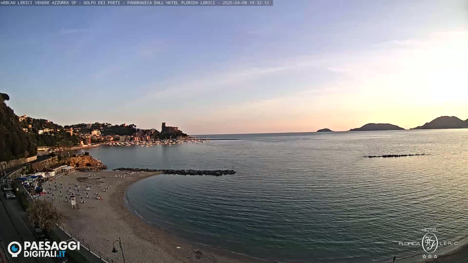 A sandy beach with lounge chairs curves into a calm, blue-green sea at sunset, backed by a town nestled against hills, under a clear, pale sky.