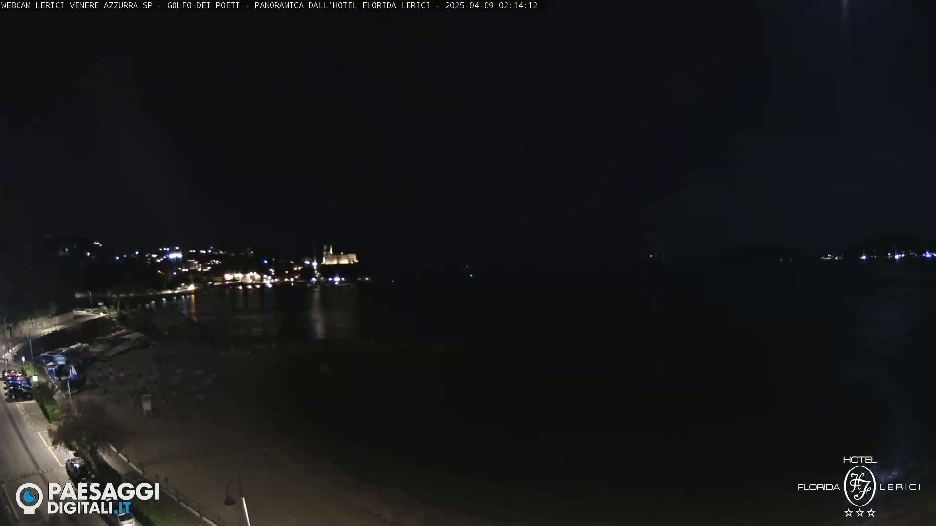 A nighttime coastal scene shows a dark body of water with a dimly lit town across it, a beach in the foreground, and a road with a few vehicles.