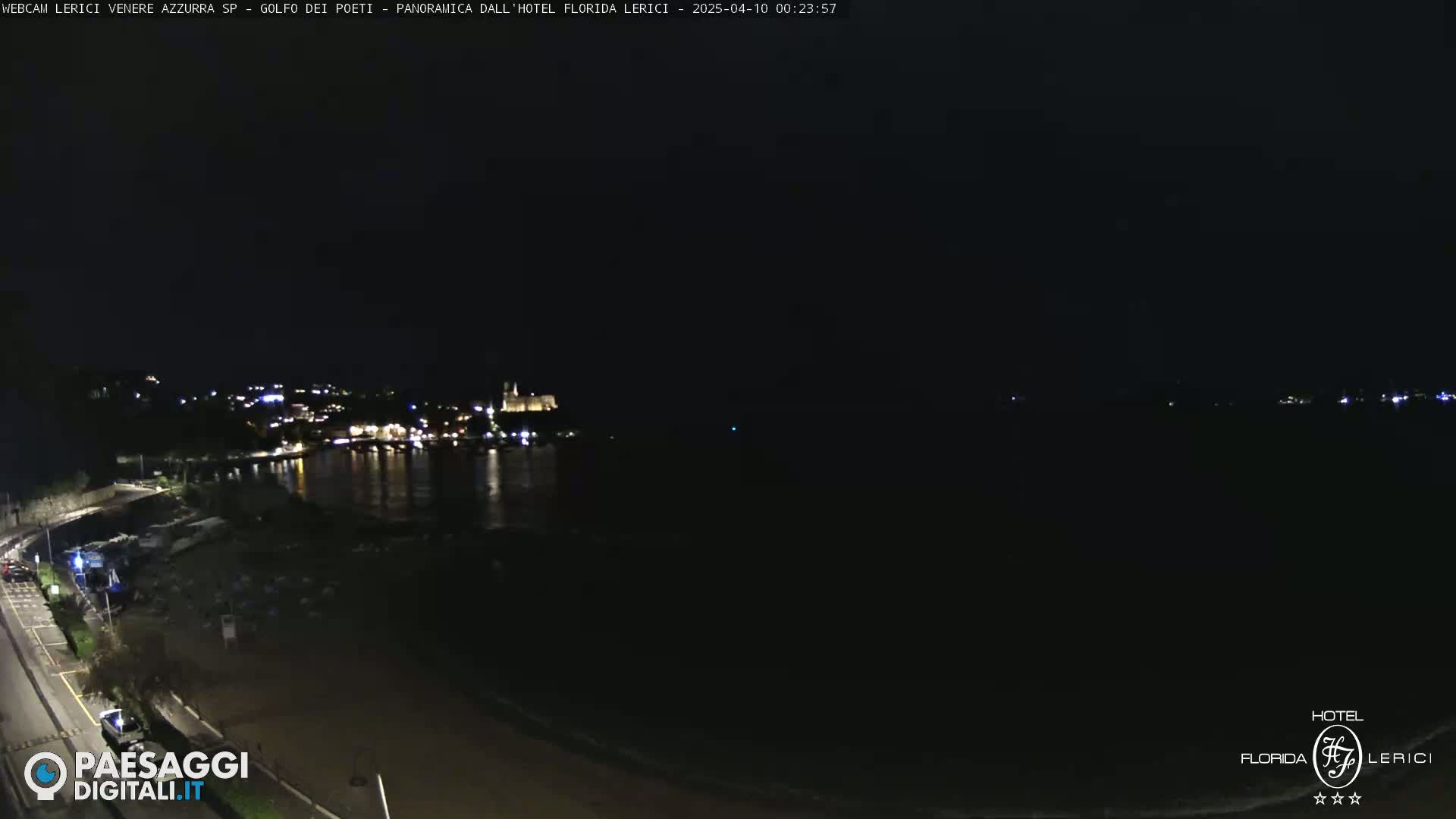 A nighttime, seaside view shows a dark body of water reflecting the lights of a distant town.