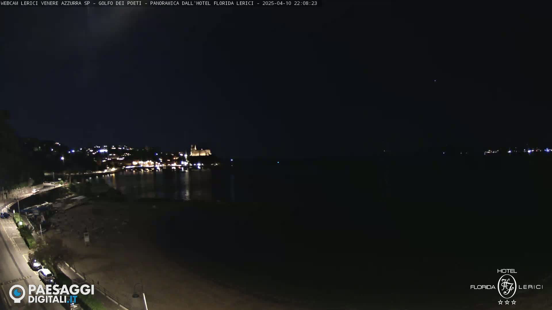 A nighttime, coastal scene shows a dark beach, a calm body of water reflecting the lights of a town across the water, and a building illuminated in the distance.