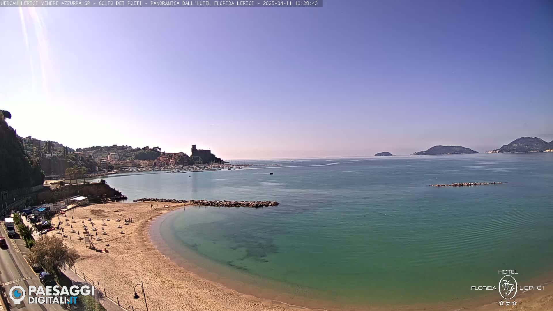 A sandy beach with a few people on it curves into a calm, teal sea, with a town and islands visible in the distance under a clear sky.