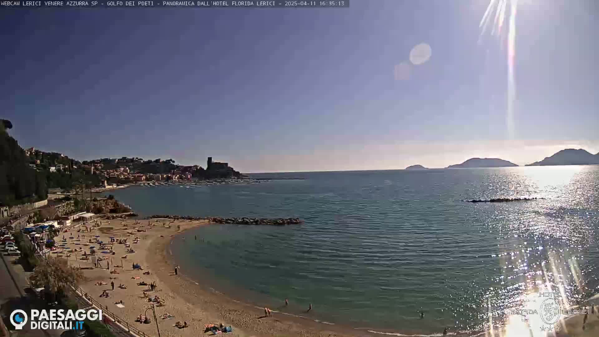 A sunny beach with many people on it curves into a calm bay, which extends to a larger body of water with distant hills under a bright, clear sky.