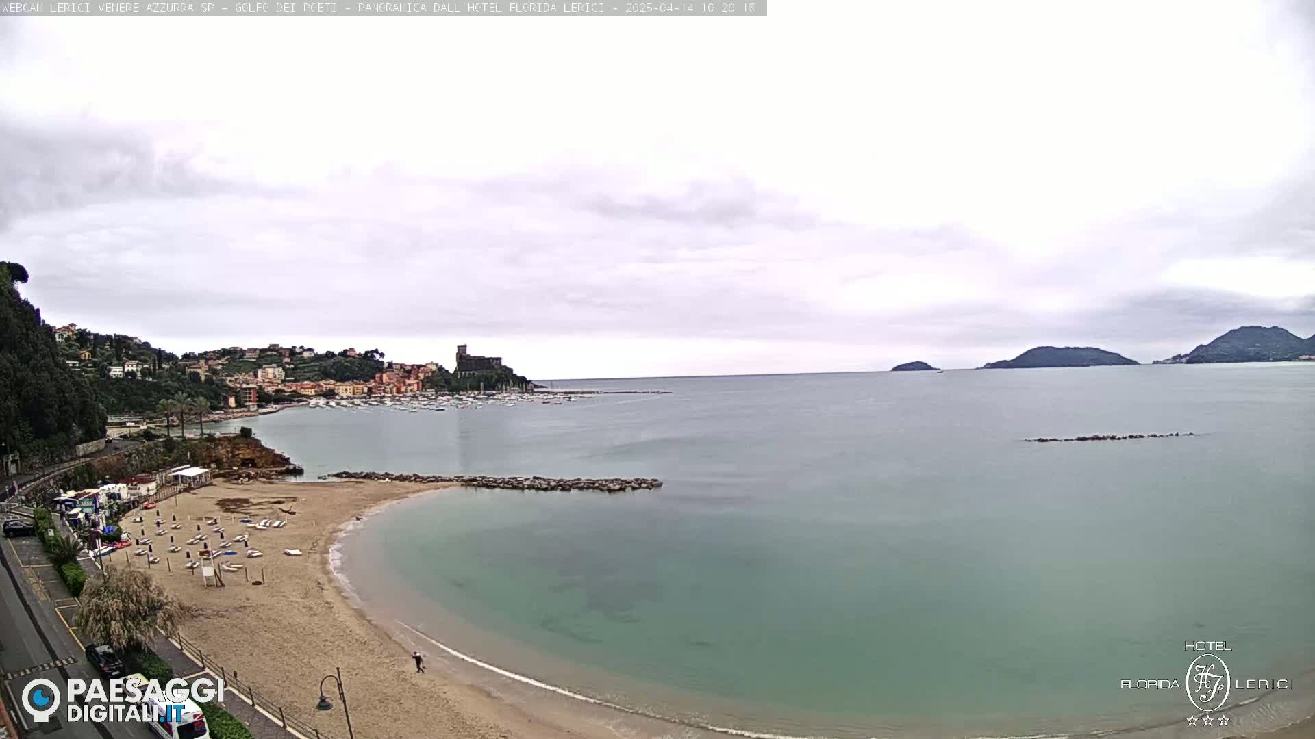 A sandy beach with calm, teal water curves around a rocky jetty, overlooking a harbor filled with boats and a town nestled against hills under an overcast sky.