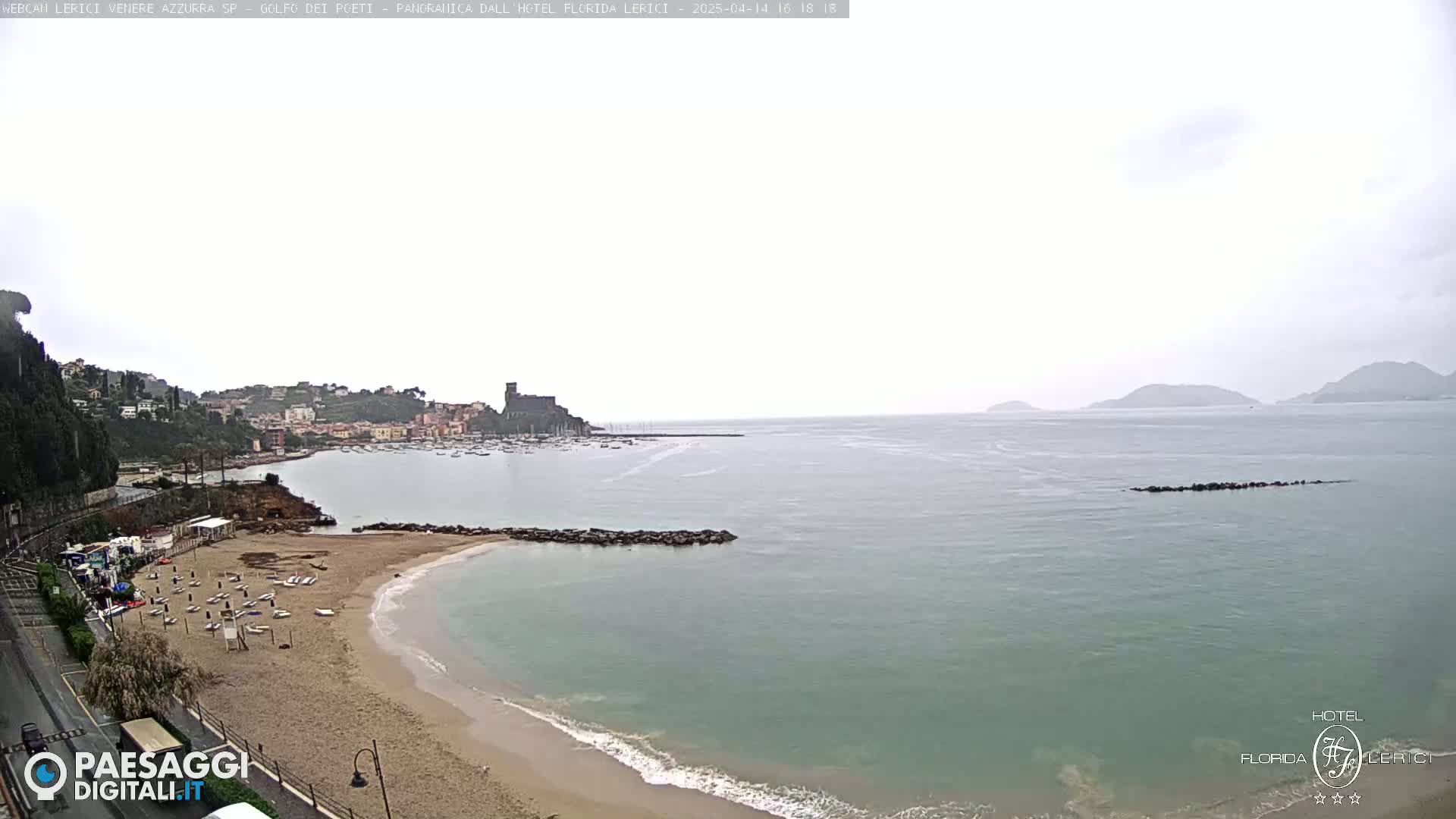 A sandy beach with a few people and boats on calm, light-grey water next to a town nestled on a hillside, under an overcast sky.