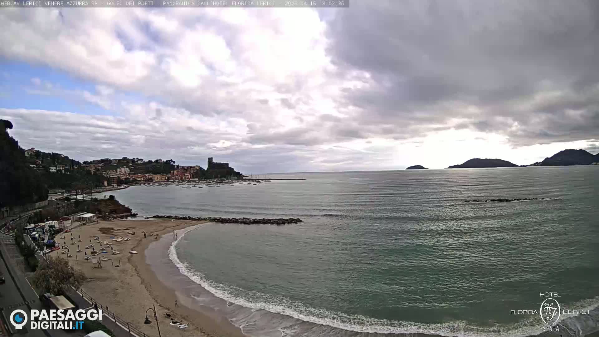 A mostly cloudy day reveals a beach with small waves lapping the shore, a calm bay with boats, and hills in the distance.