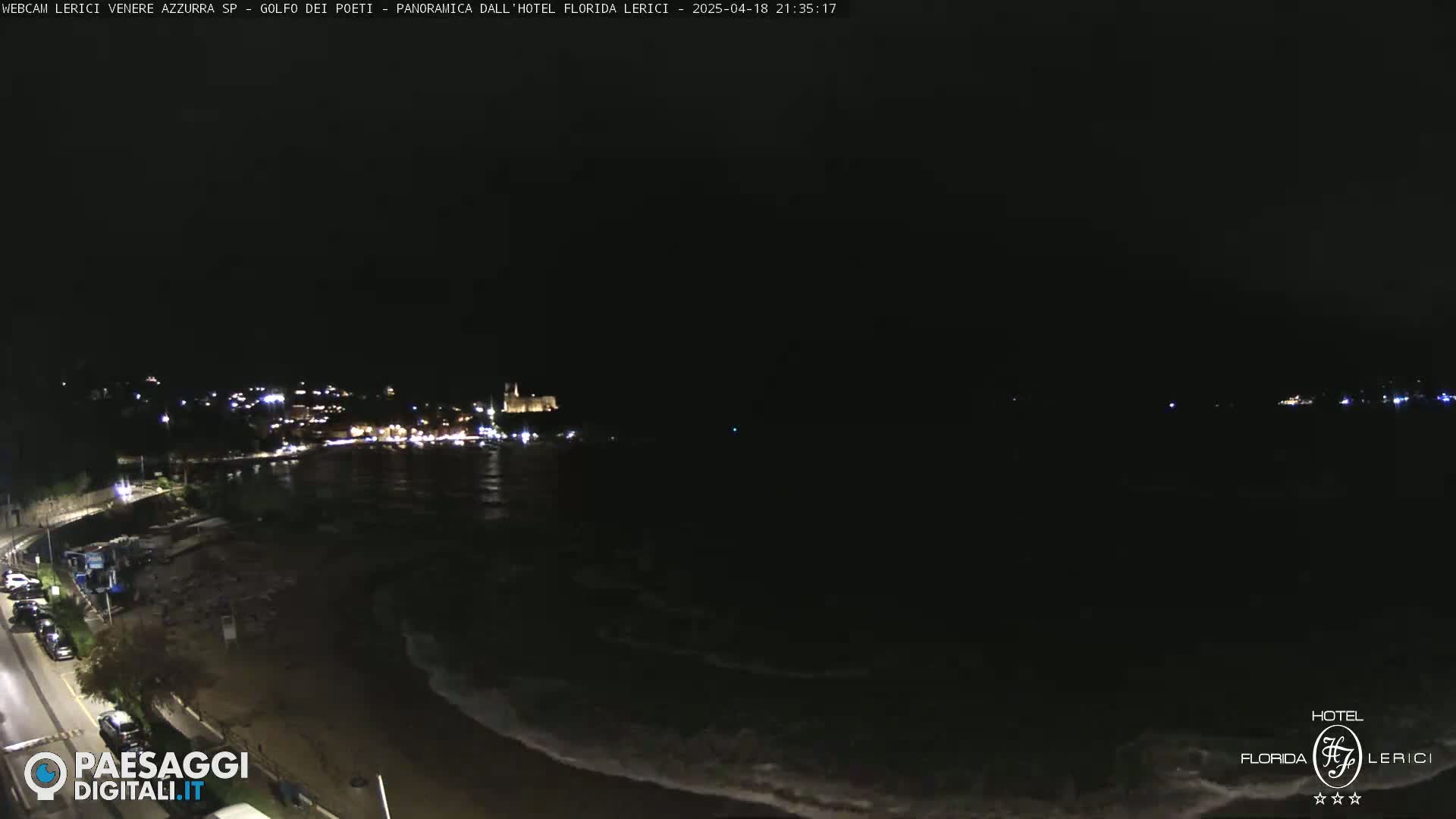 A dark nighttime coastal scene shows a town's lights across a body of water with gentle waves lapping at a shore.