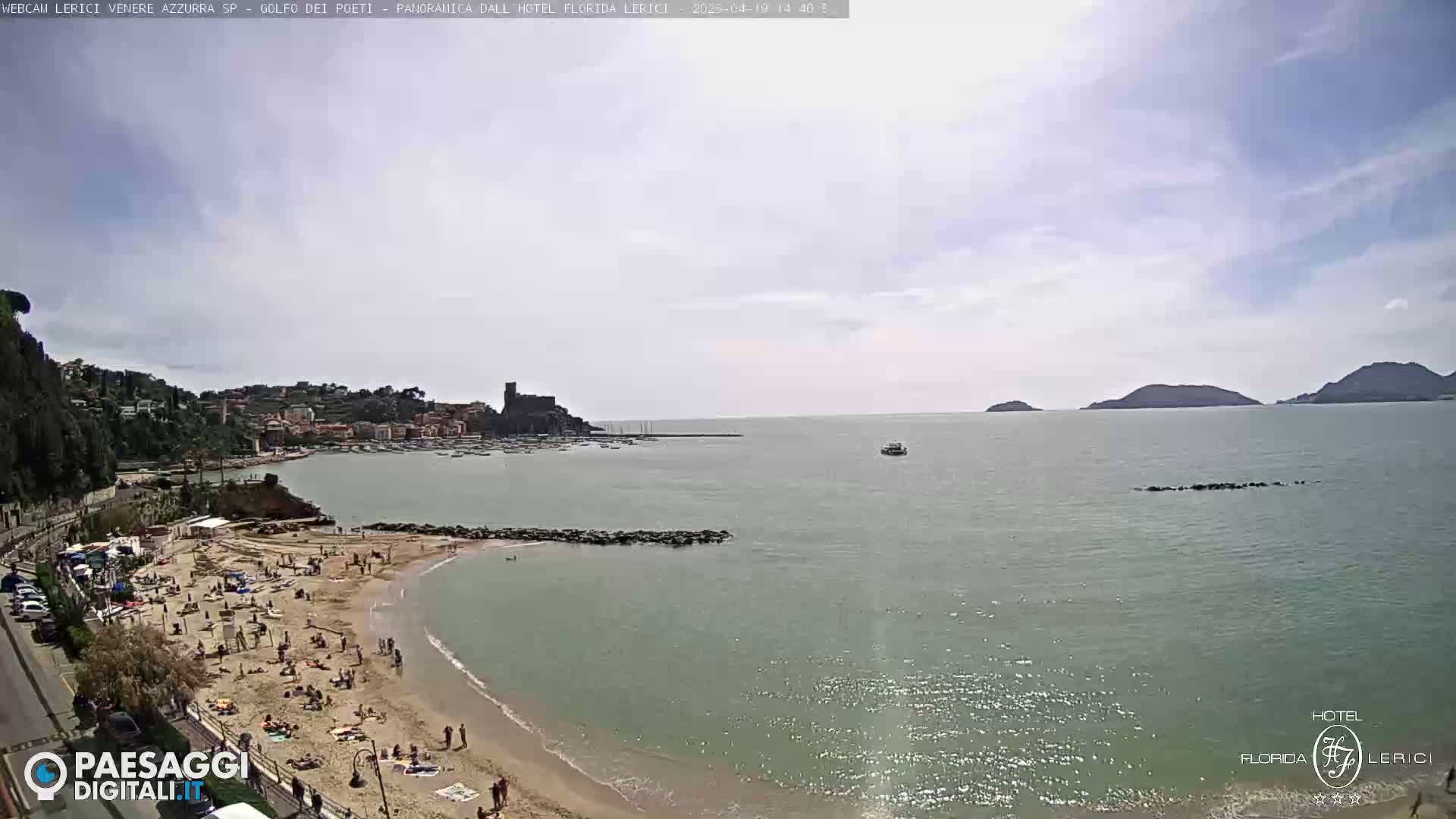 A mostly sunny day shows a sandy beach crowded with people, a calm bay with a town and castle in the background, and islands visible in the distance.