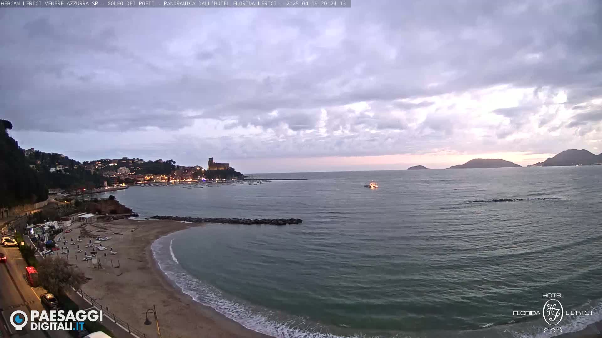 A mostly cloudy twilight scene shows a calm bay with a sandy beach, a town nestled on a hillside, and distant islands under a pale sky.