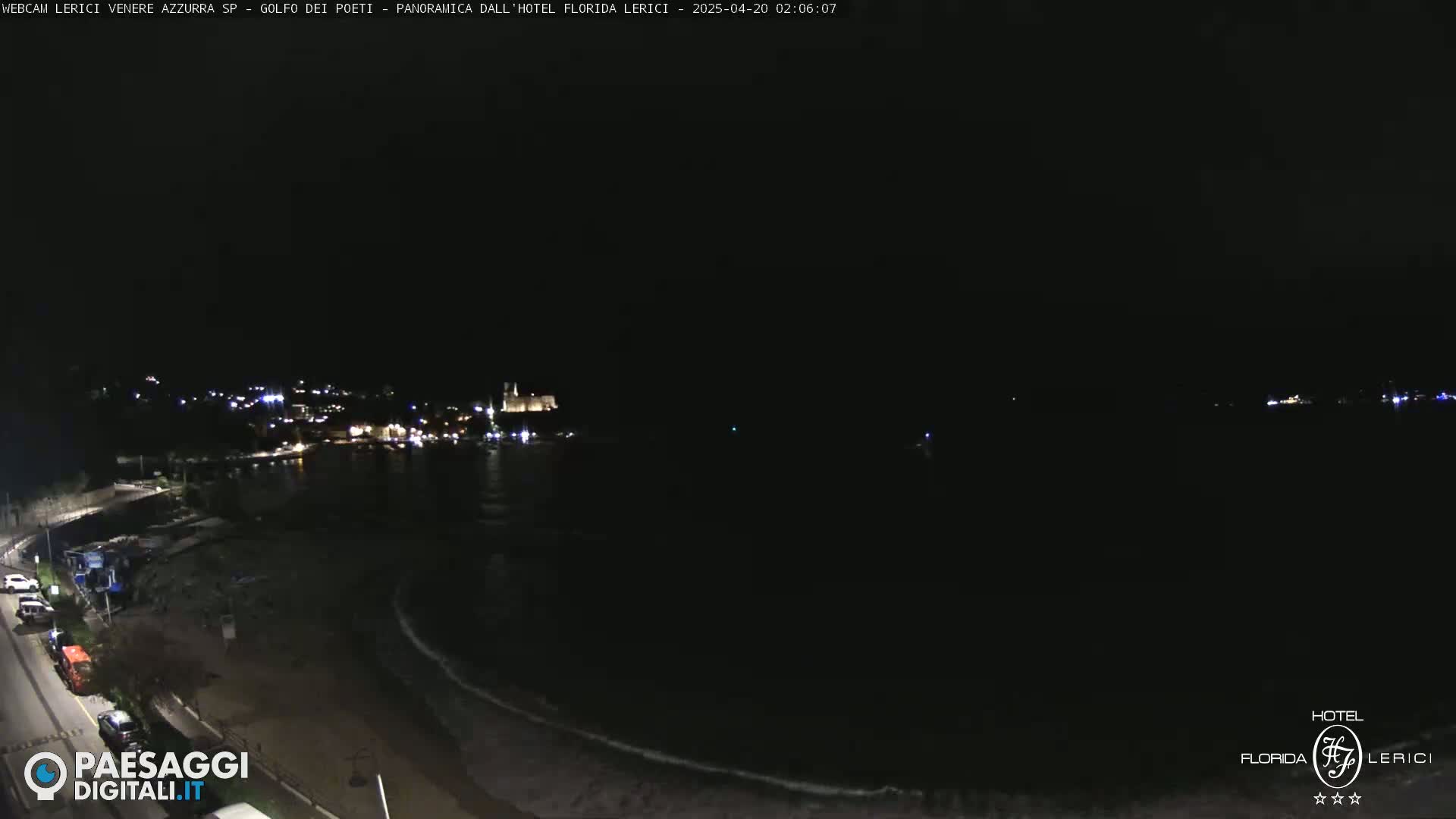 A nighttime view of a dark coastline with a few lights visible in the distance, showing a calm, dark sea.