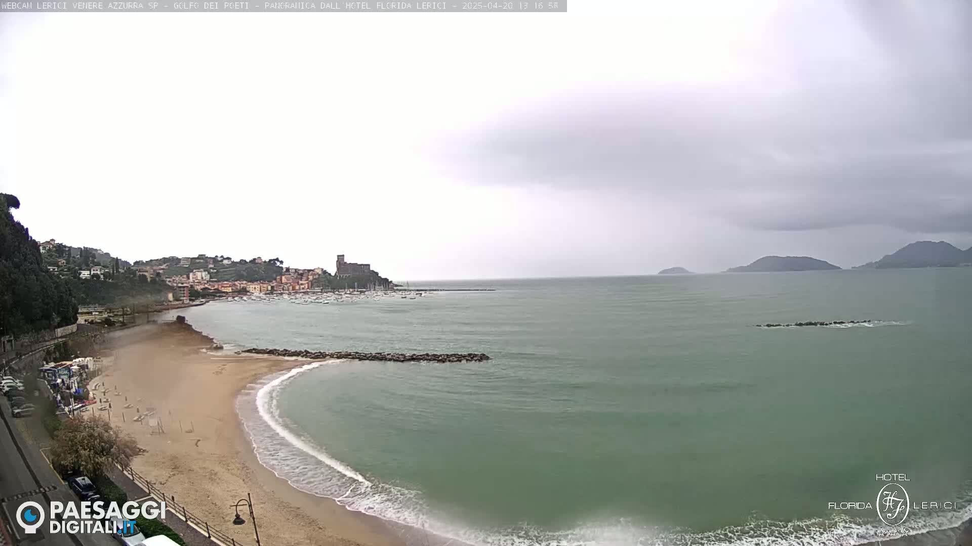 A sandy beach curves into a calm, greenish sea under an overcast sky, with a town and hills visible in the distance.