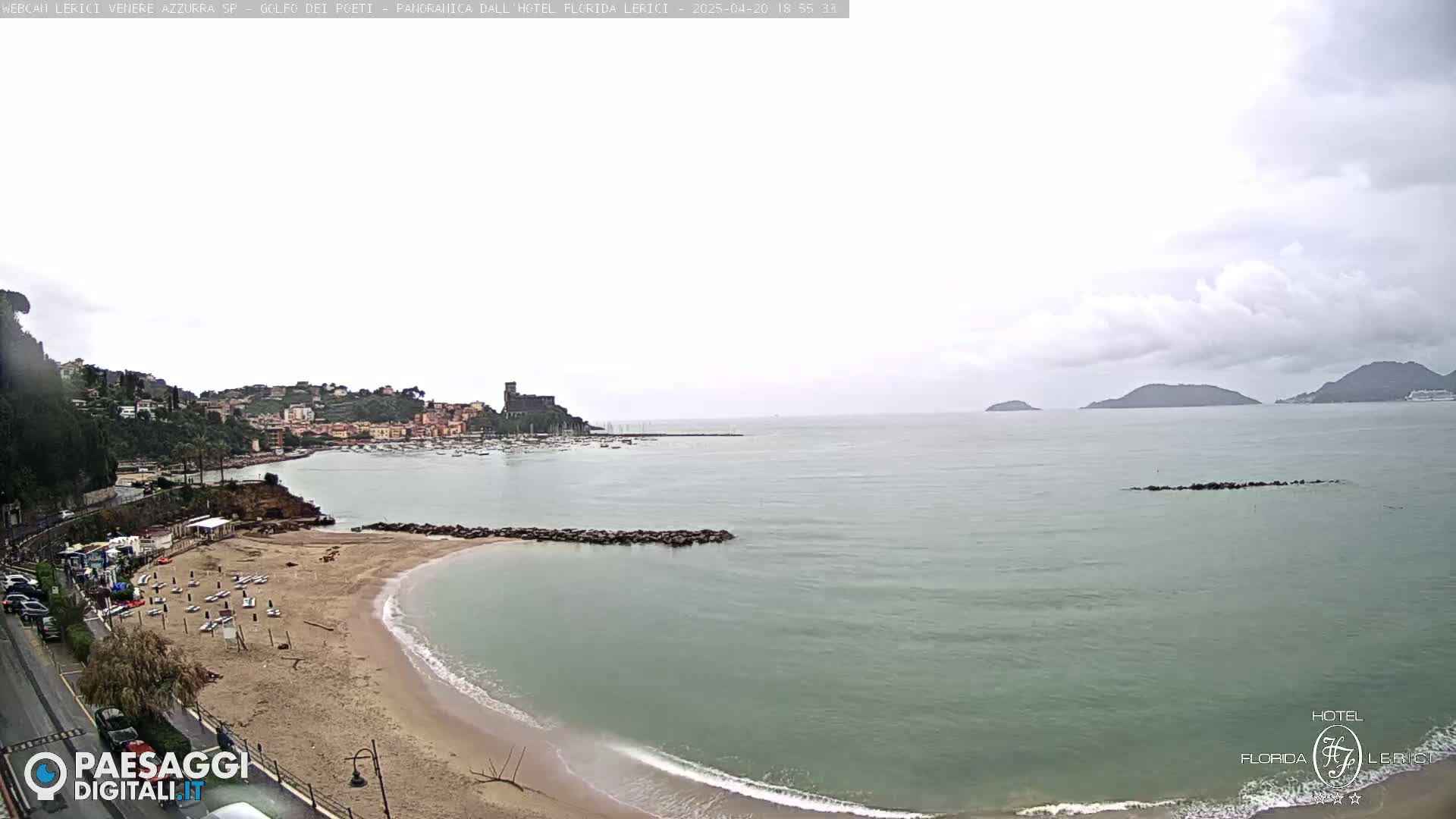 A partly cloudy day shows a sandy beach curving into a calm bay, with a town and hills in the background, and islands visible on the horizon.
