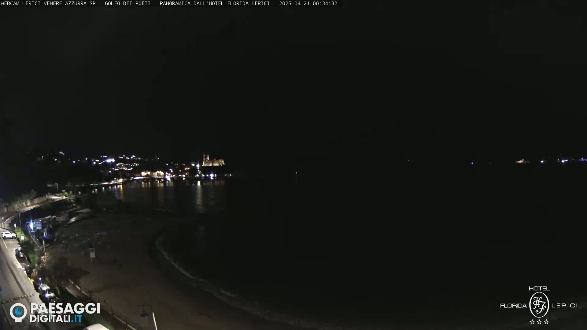 A nighttime view of a dark, sandy beach and calm ocean, with a town’s lights visible across the water.