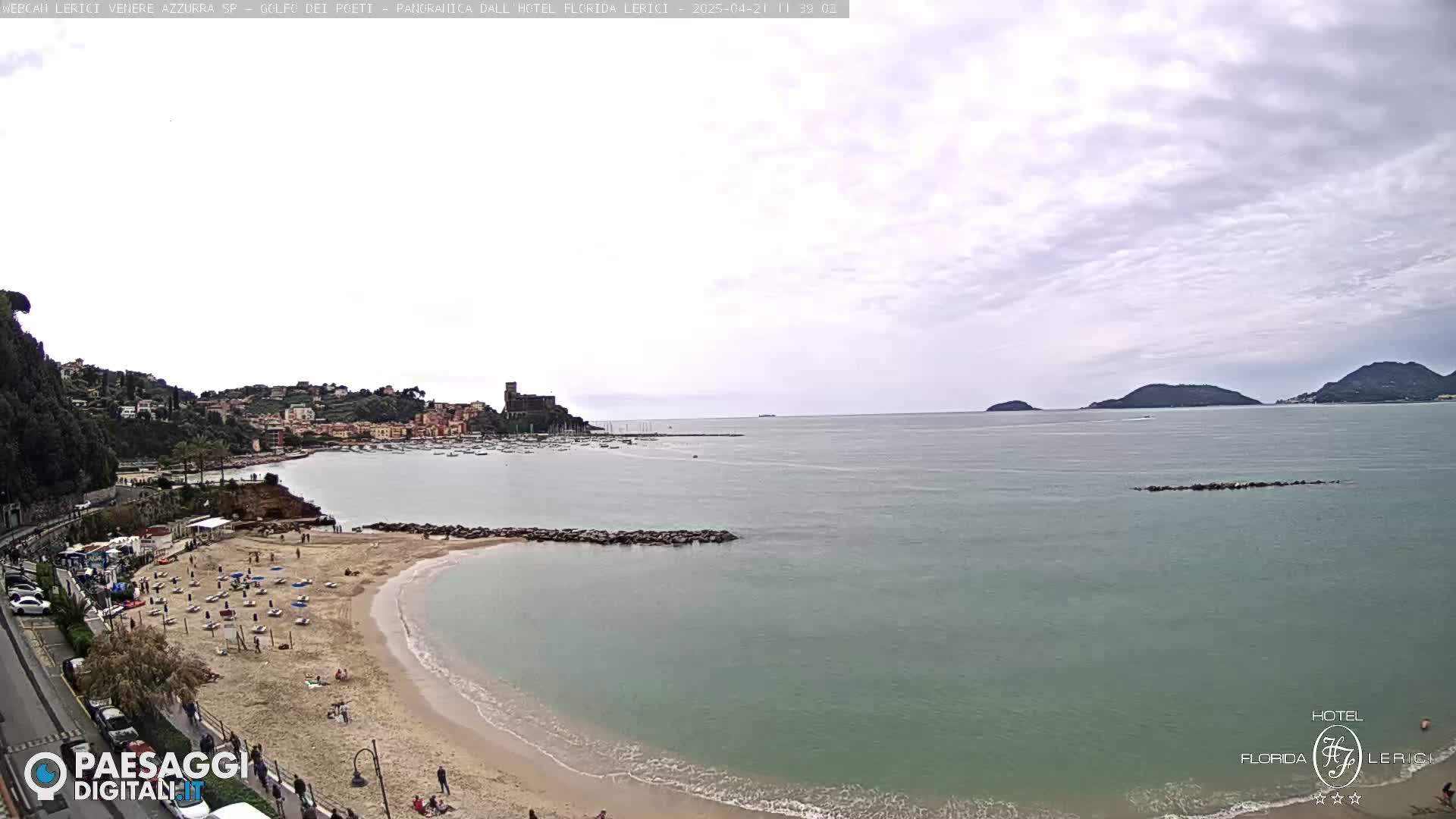 A sandy beach with people on it curves into a calm, greenish-blue sea under a mostly cloudy sky, with a town and distant islands visible in the background.