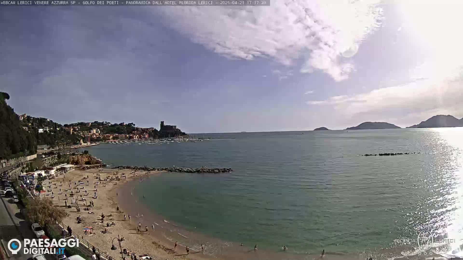 A partly sunny day shows a sandy beach with people sunbathing and swimming, next to a calm sea with boats and distant hills under a partly cloudy sky.