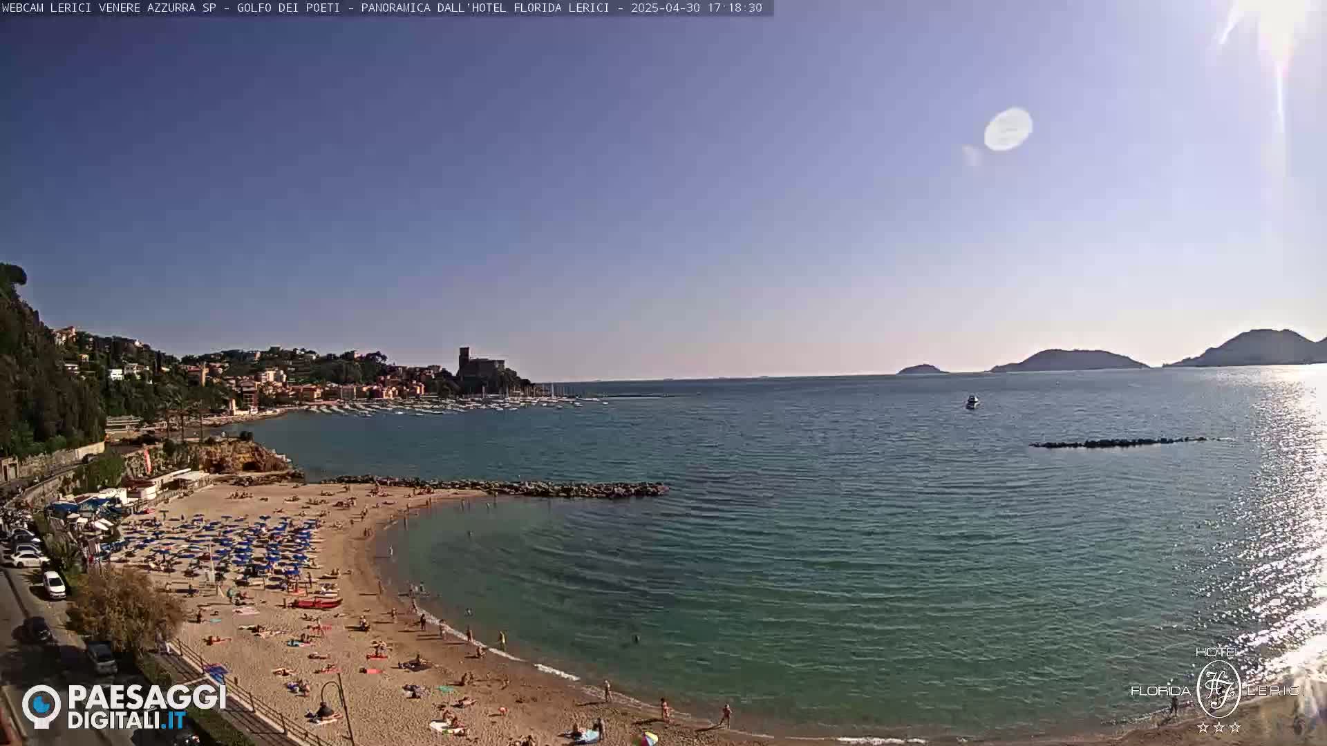 A sunny day at a sandy beach cove with clear turquoise water, numerous beach umbrellas, and bathers, overlooks a calm bay with boats and distant hills.