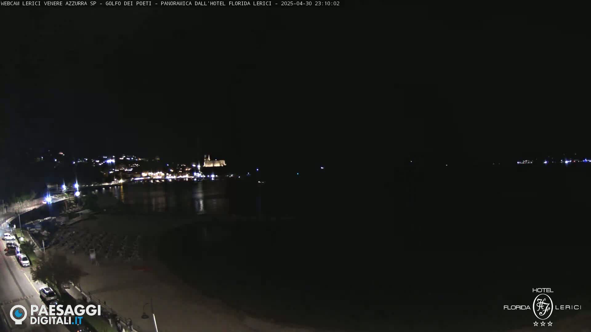 A nighttime view of a dark, calm body of water, a shoreline with sparsely lit buildings and parked cars, and a dark beach with some beach chairs.