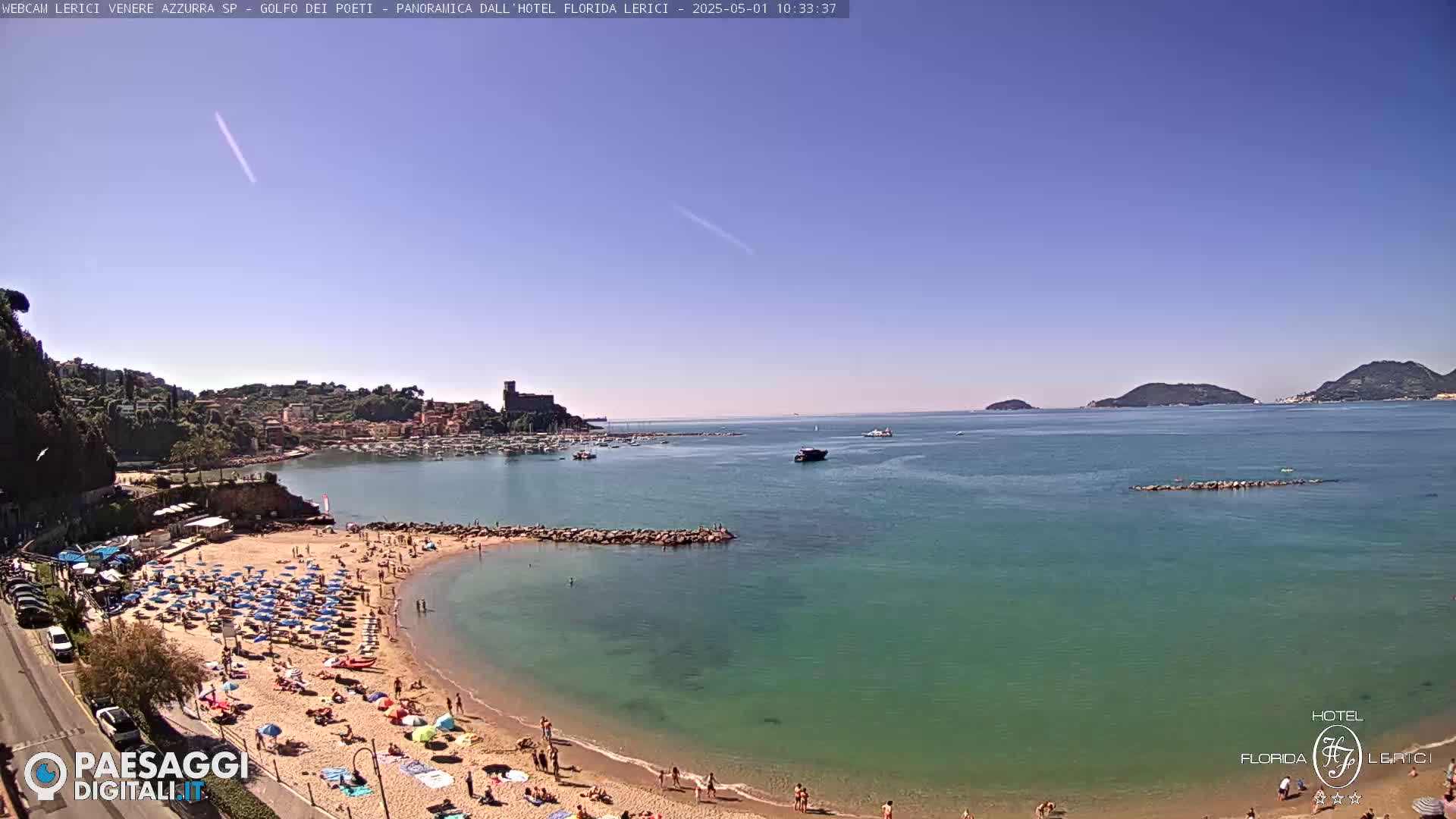A sunny day shows a crowded beach curving into a calm, teal sea, with boats in a bay and islands in the distance.