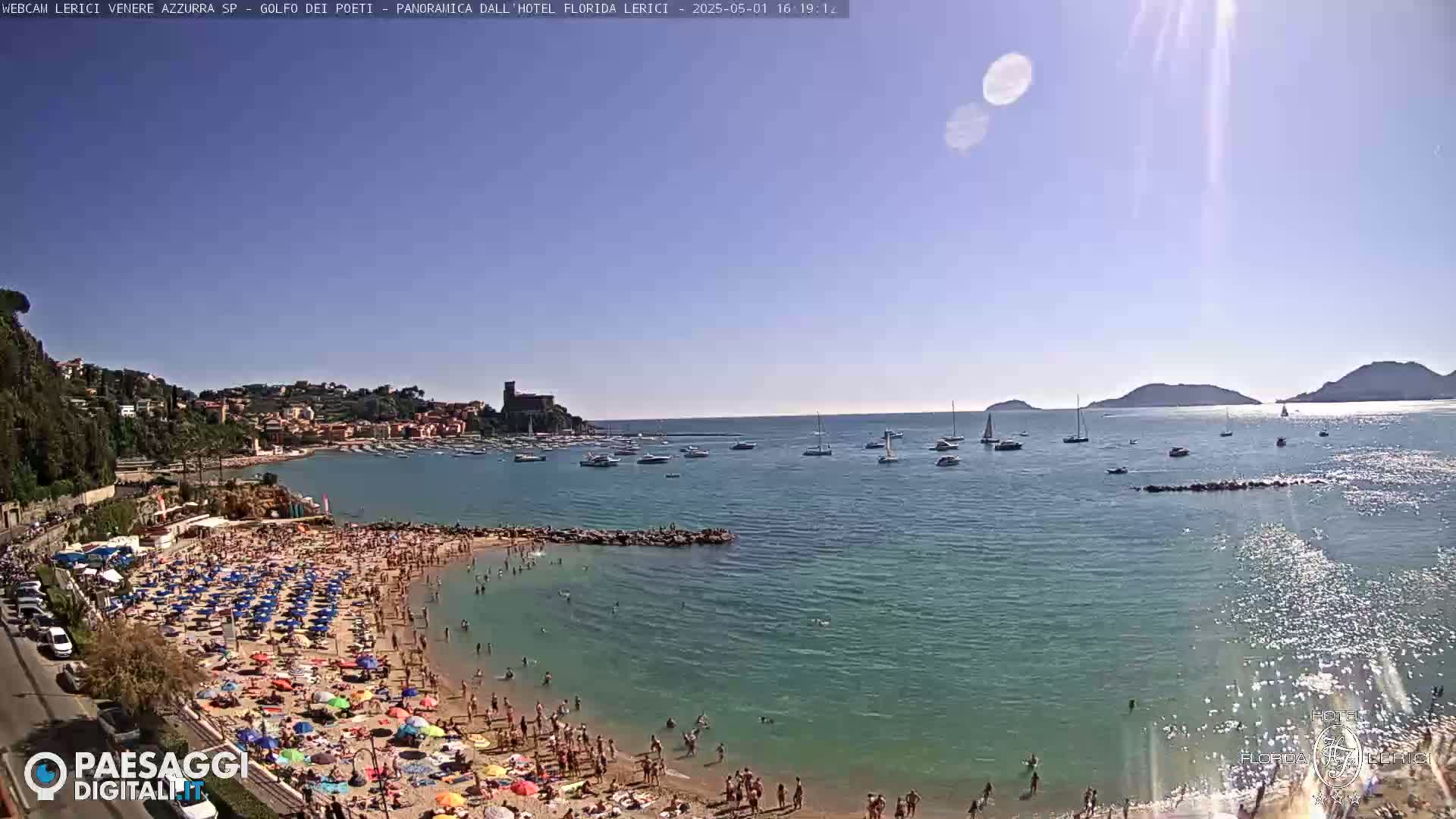 A sunny beach scene shows a crowded sandy beach with numerous colorful umbrellas, a calm bay with boats, and hills in the background.