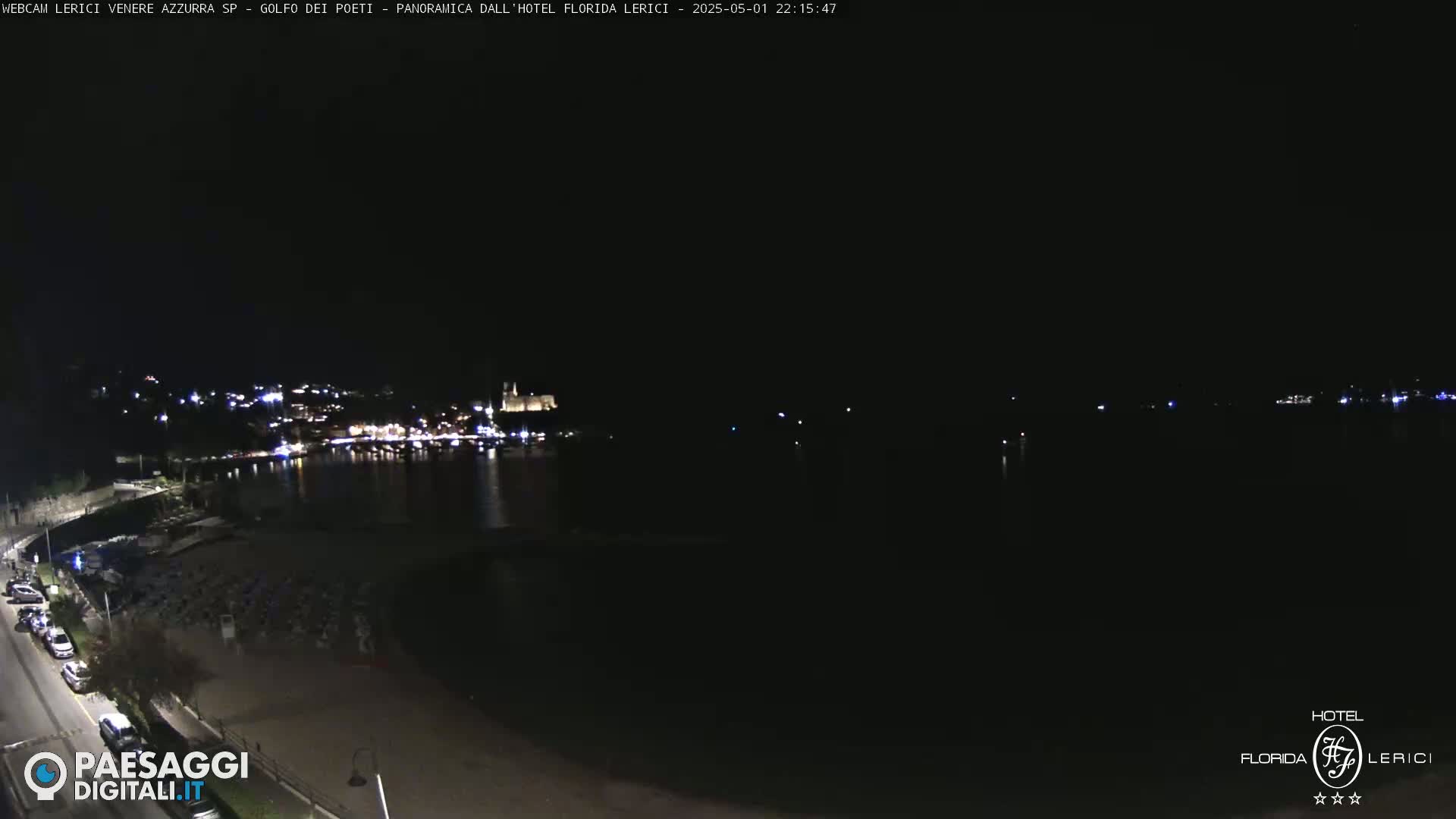 A nighttime, seaside view shows a dark beach, a road with parked cars, and a town across the water illuminated with lights.