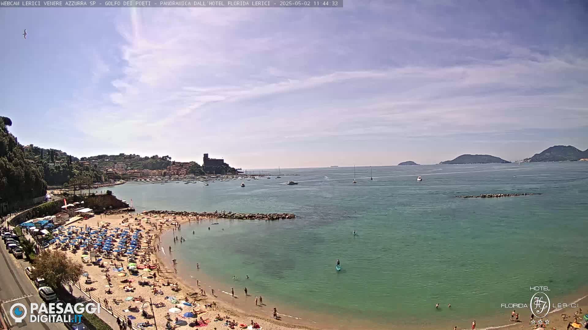 A sunny day at a crowded beach with calm, clear turquoise water, boats in the bay, and hills in the distance.