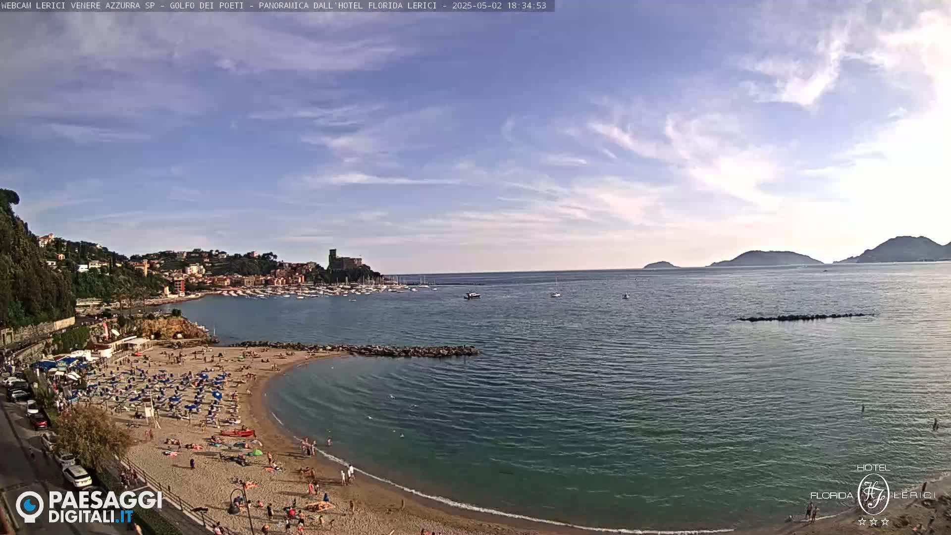 A sunny day shows a crowded sandy beach curving into a calm bay, with a town and hills rising behind it, and islands visible across the water.