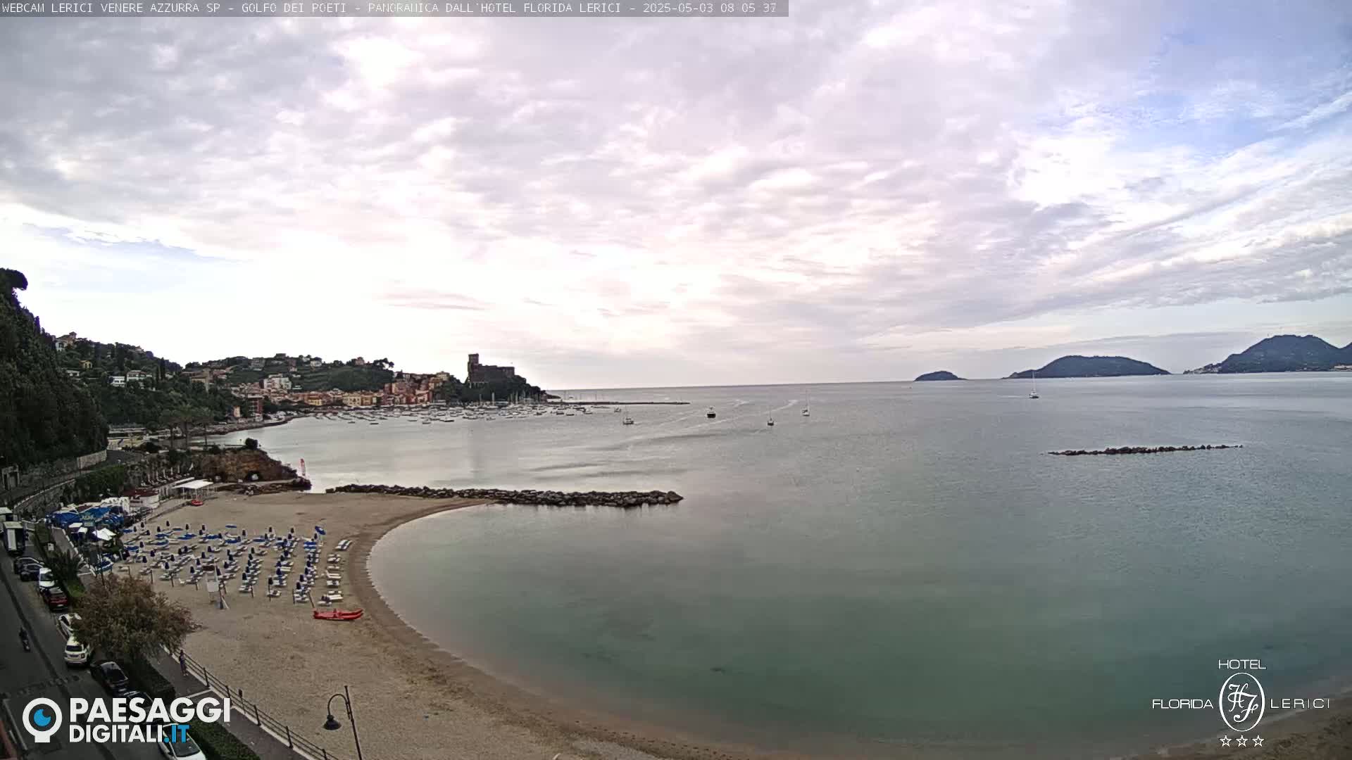 A mostly cloudy day overlooks a sandy beach with rows of lounge chairs next to calm, teal water, a town nestled on a hillside, and several boats in the bay.