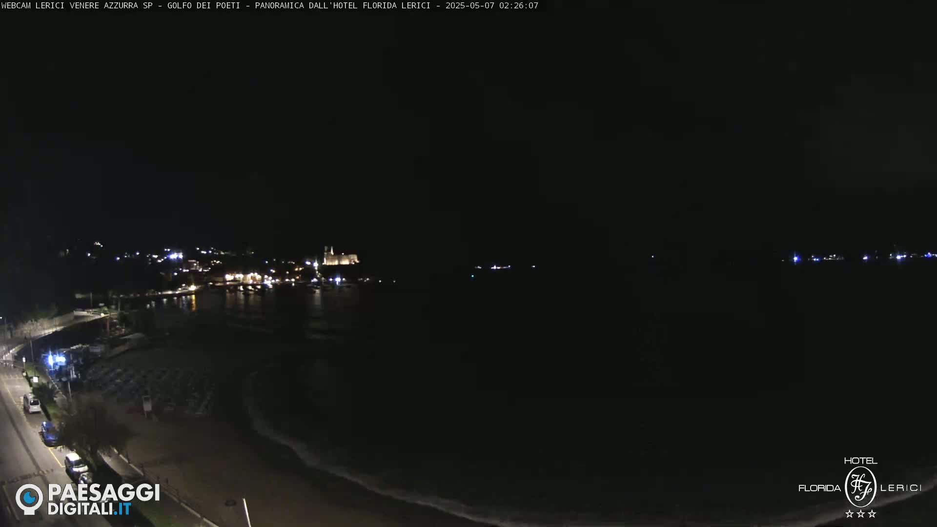 A nighttime coastal scene shows a dark beach, a calm sea, and a dimly lit town in the distance.