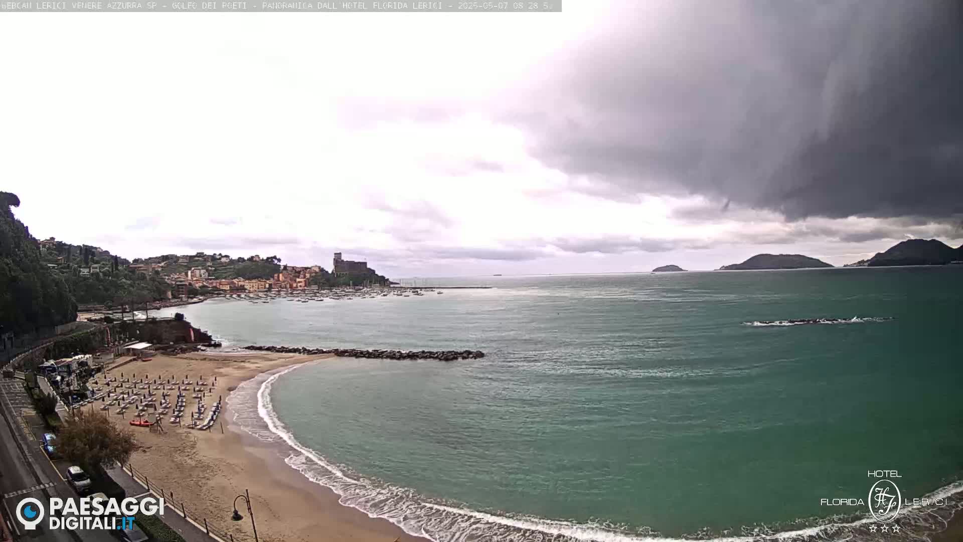 A coastal town with a sandy beach and calm, teal water under a cloudy sky.