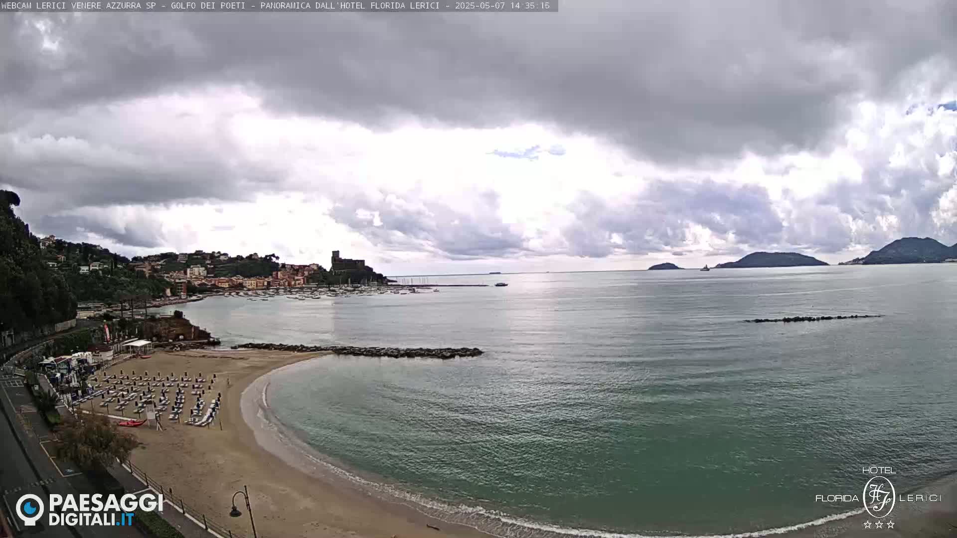 A sandy beach with rows of lounge chairs curves into a calm, teal-colored sea under a mostly cloudy sky, with hills and a town visible in the distance.