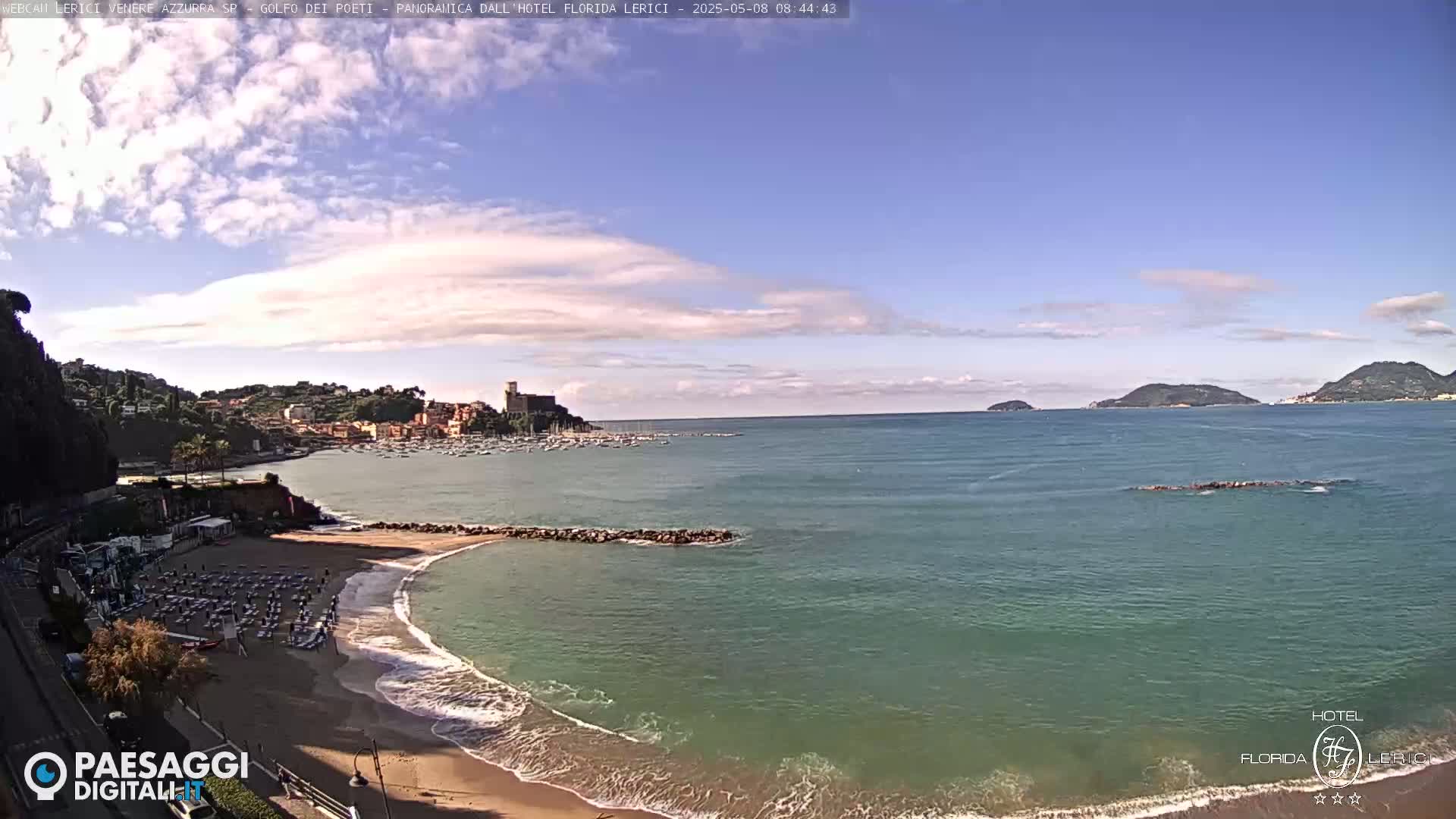 A mostly sunny day shows a sandy beach with calm, teal-colored water lapping the shore, backed by a town with buildings and a small harbor, and distant islands under a mostly clear sky.