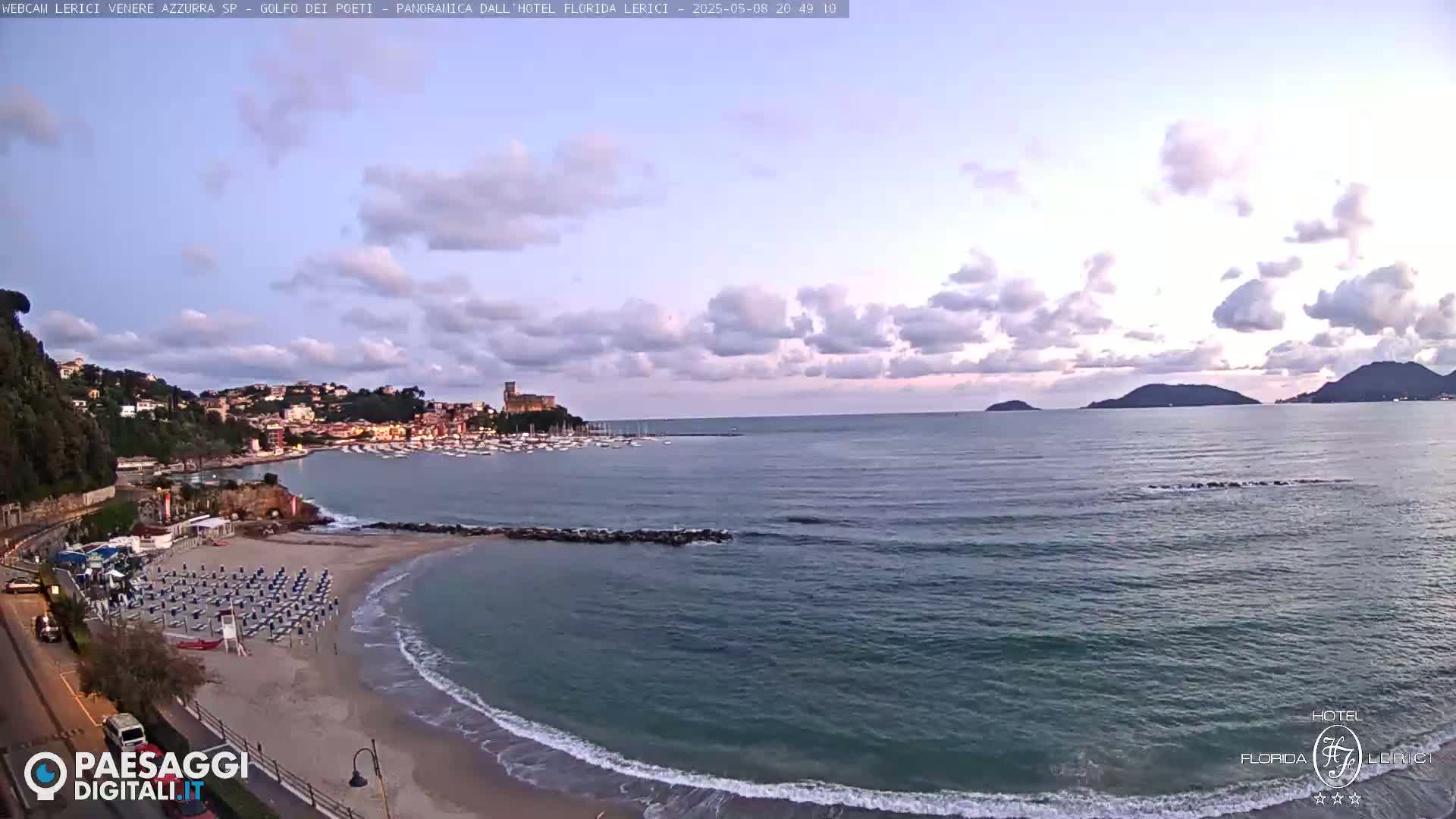 A partly cloudy evening scene shows a sandy beach with rows of beach umbrellas and chairs next to a calm sea with boats in a bay and hills in the distance.