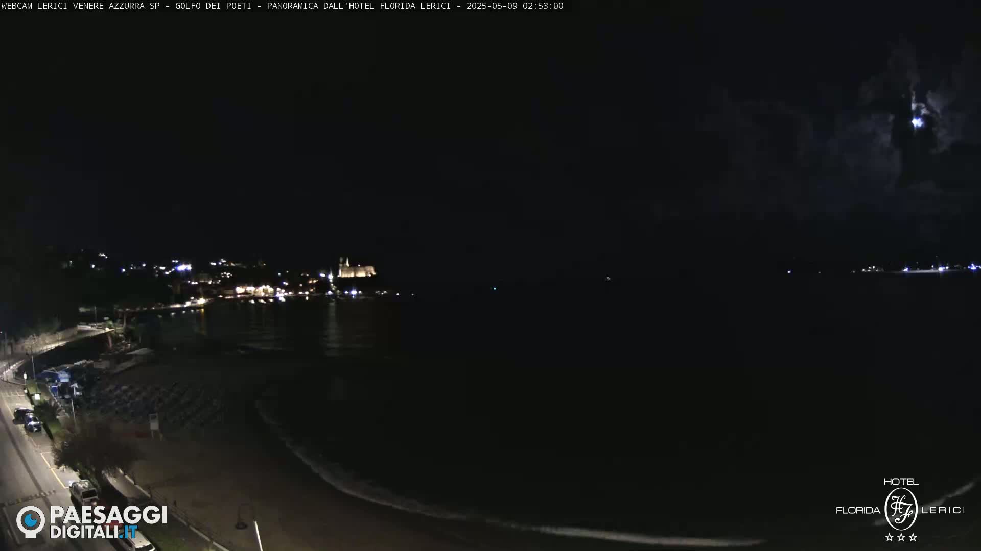 A nighttime coastal scene shows a dark body of water lapping a beach with a town's lights visible in the distance under a partly cloudy sky.