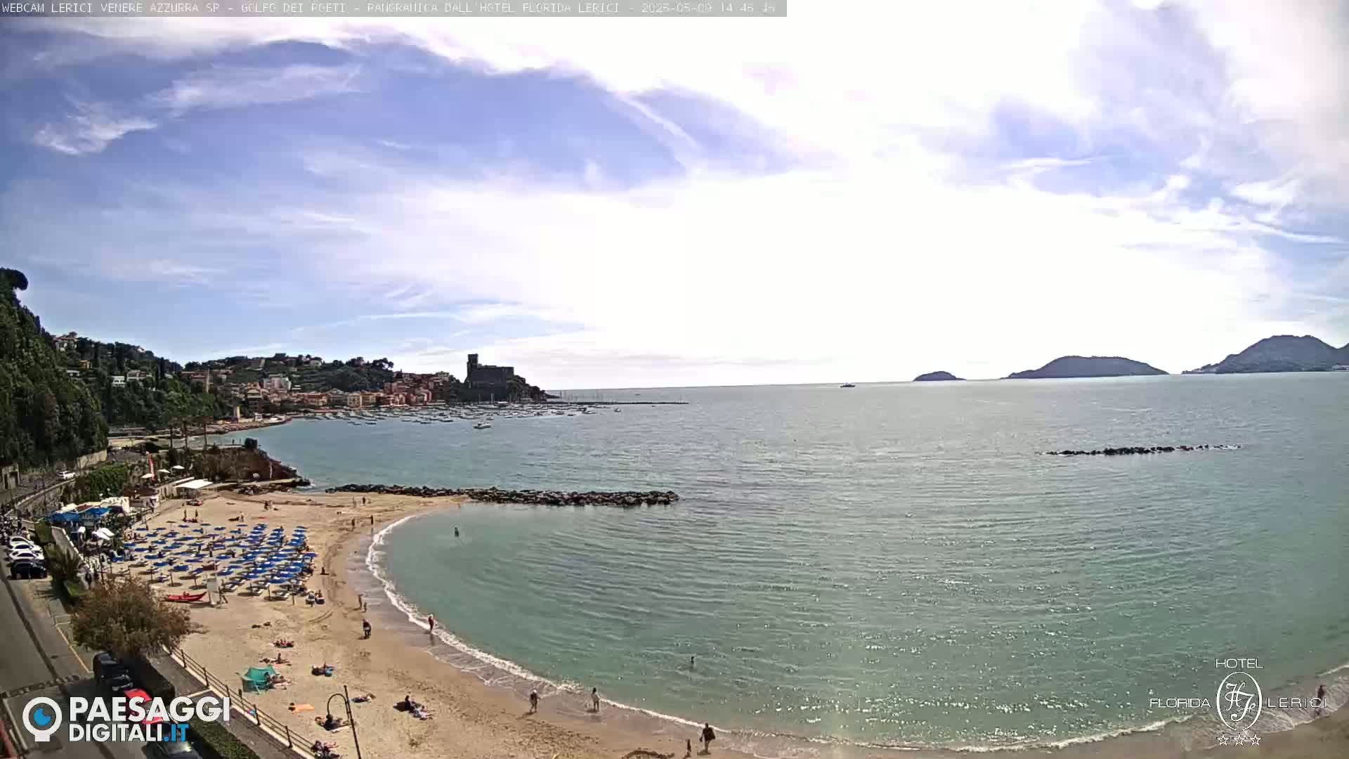 A sandy beach with numerous blue umbrellas and sunbathers is nestled beside a calm, clear bay, with a town and distant hills visible under a partly cloudy sky.