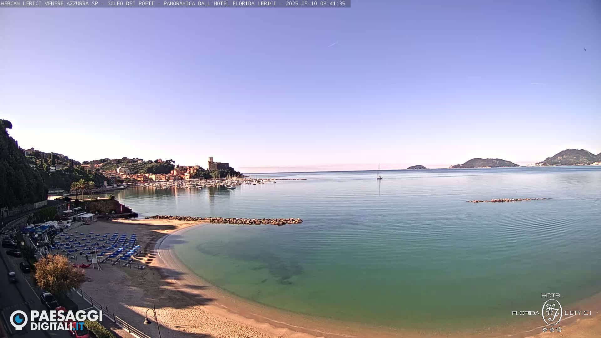 A sandy beach with blue umbrellas and calm, clear turquoise water is situated next to a small town nestled against hills, under a clear blue sky.