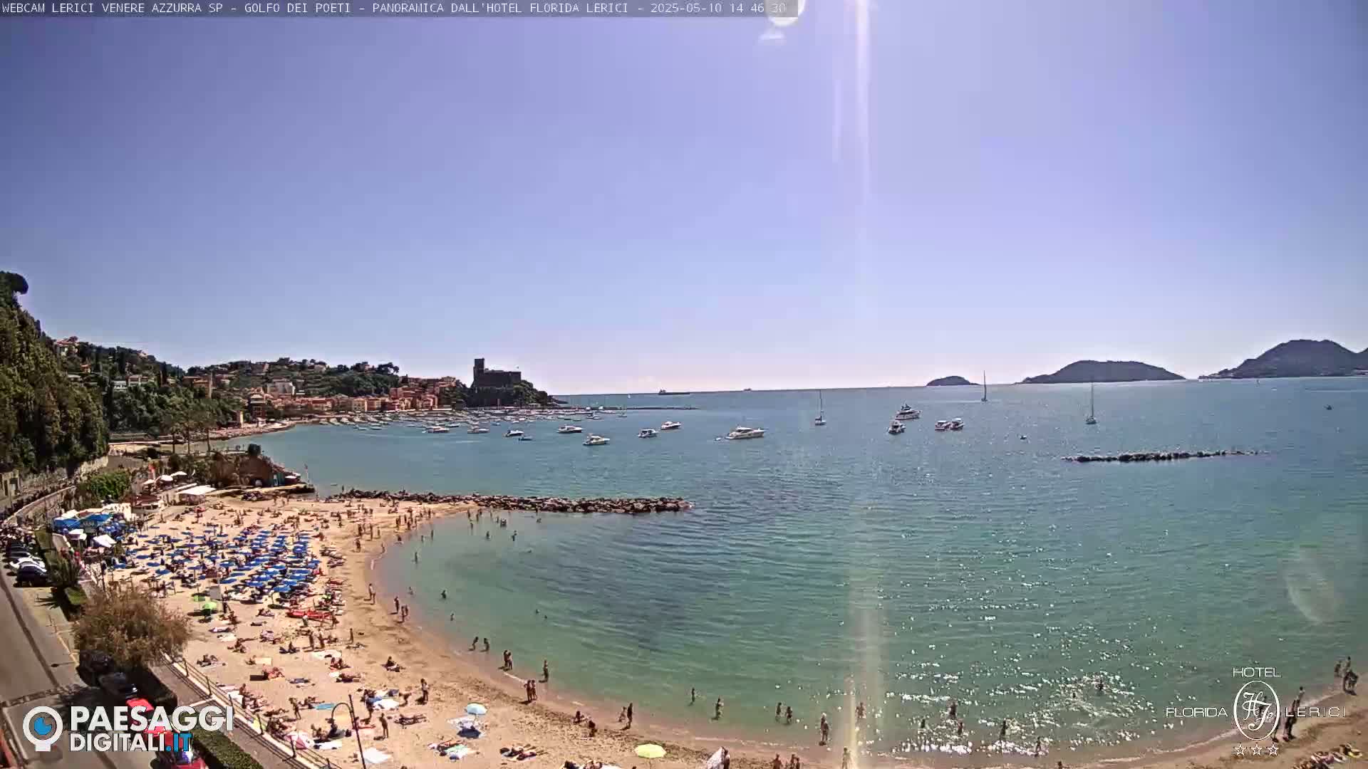 A sunny beach scene shows a sandy shore crowded with people and umbrellas, a calm, clear sea with boats, and a town nestled against a hillside in the background.