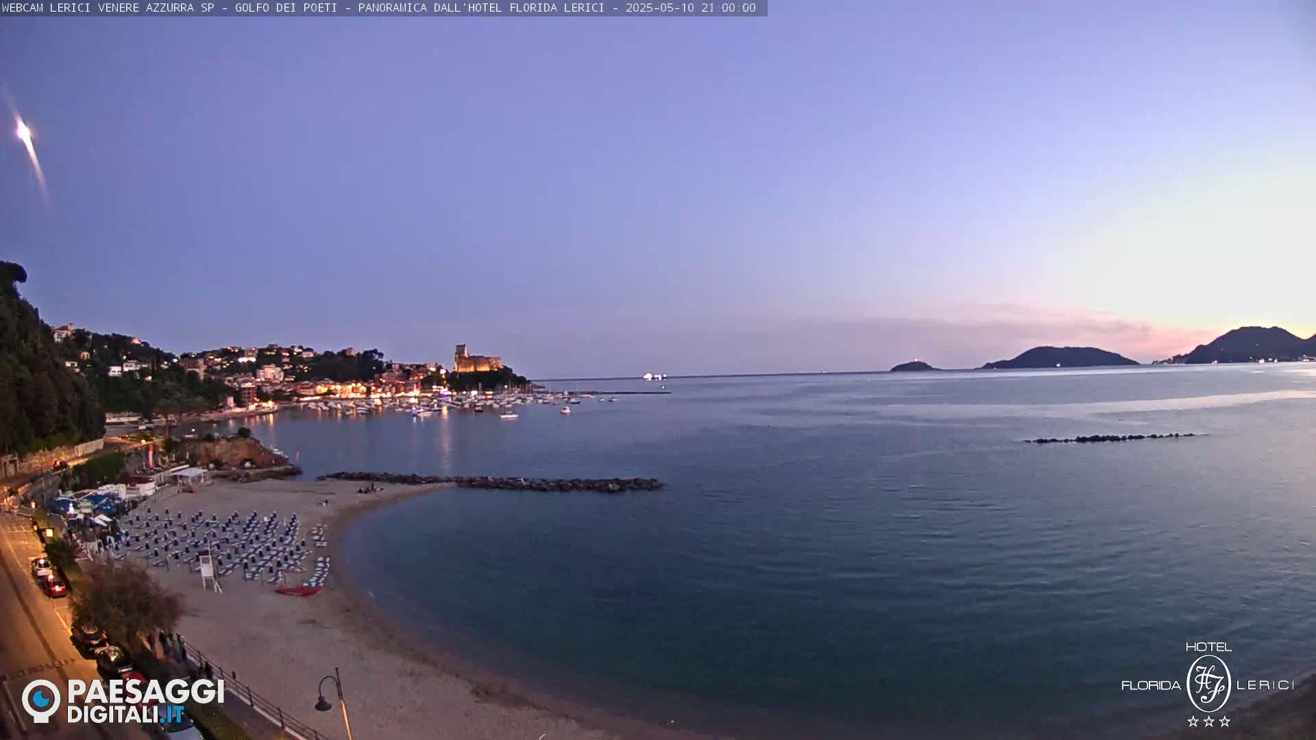 A twilight view shows a calm bay with a sandy beach, a town with lights, and distant hills under a clear, dusky sky.
