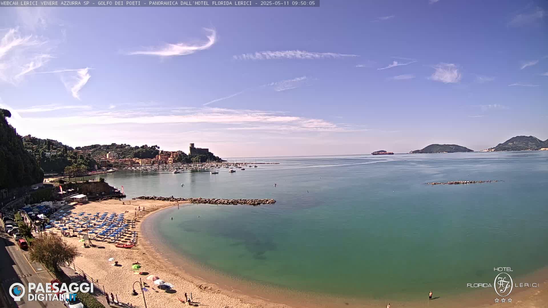 A sunny day shows a partly cloudy sky over a calm, teal-colored sea, with a sandy beach, a town nestled against a hillside, and boats in a harbor.
