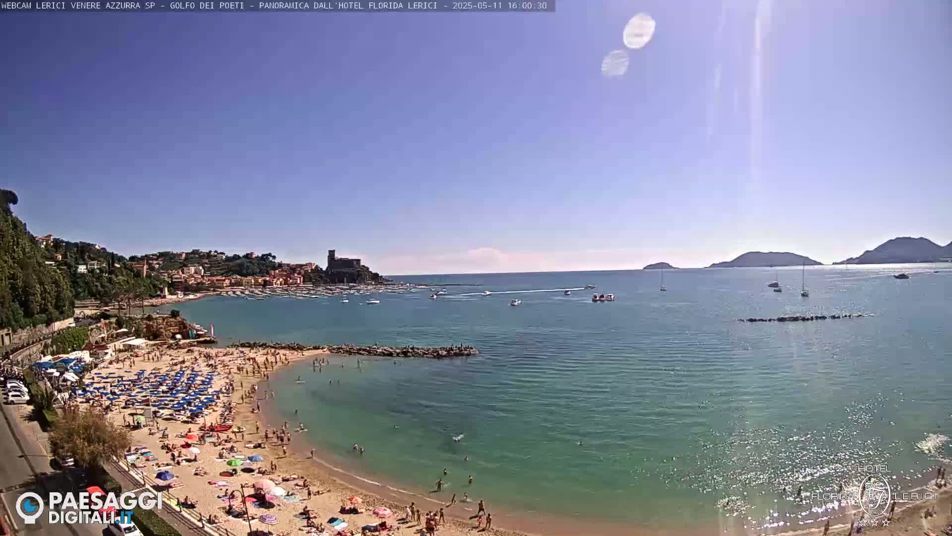 A sunny day at a crowded beach with clear, calm water, boats in the bay, and hills in the distance.