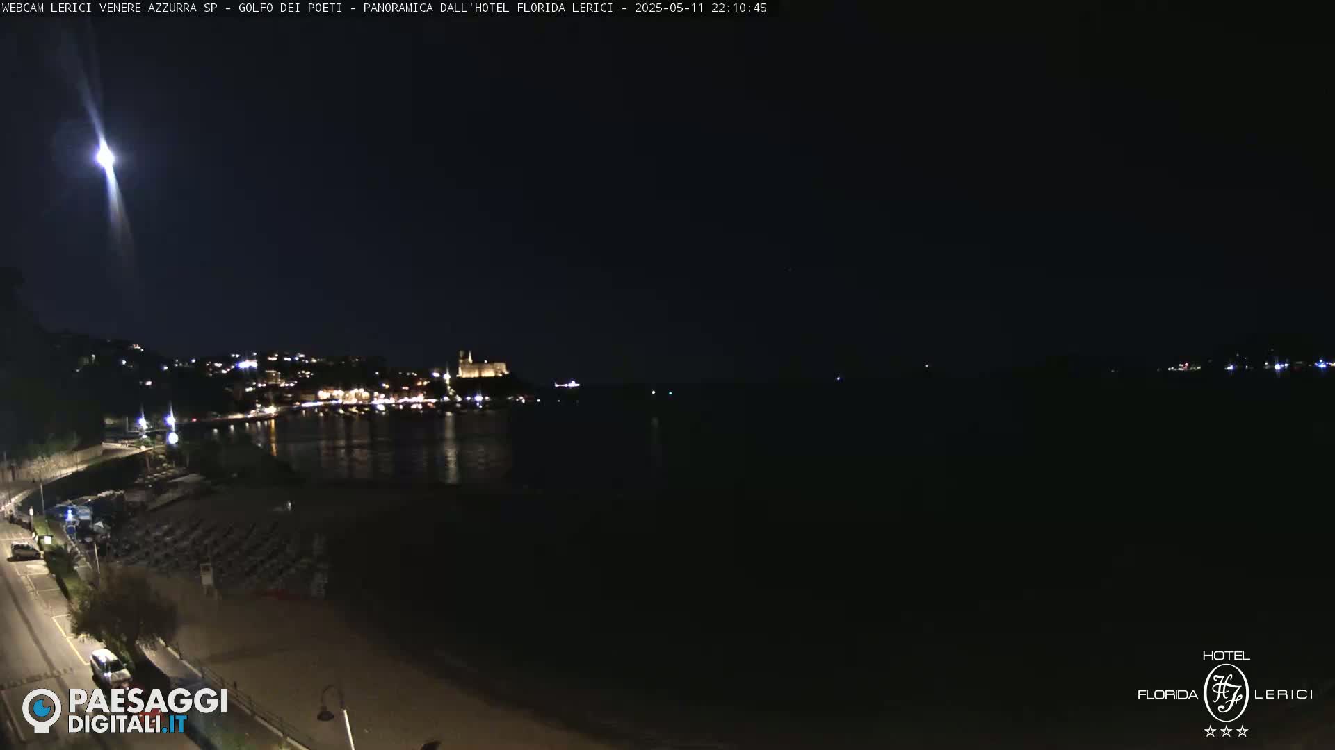 A nighttime, seaside view shows a dark beach with rows of empty beach chairs, a calm body of water reflecting the lights of a town on the opposite shore under a clear, dark sky.