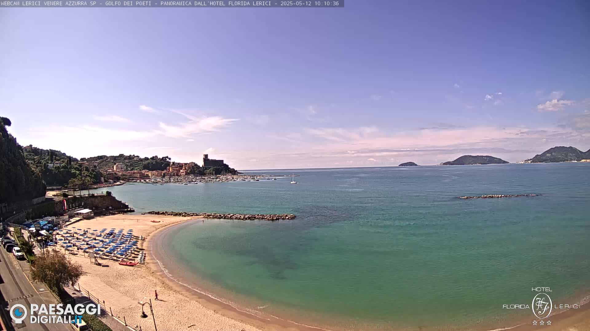 A mostly sunny day shows a sandy beach with rows of beach umbrellas and chairs, a calm teal sea, and a town nestled on a hillside in the distance, with islands visible further out on the horizon.