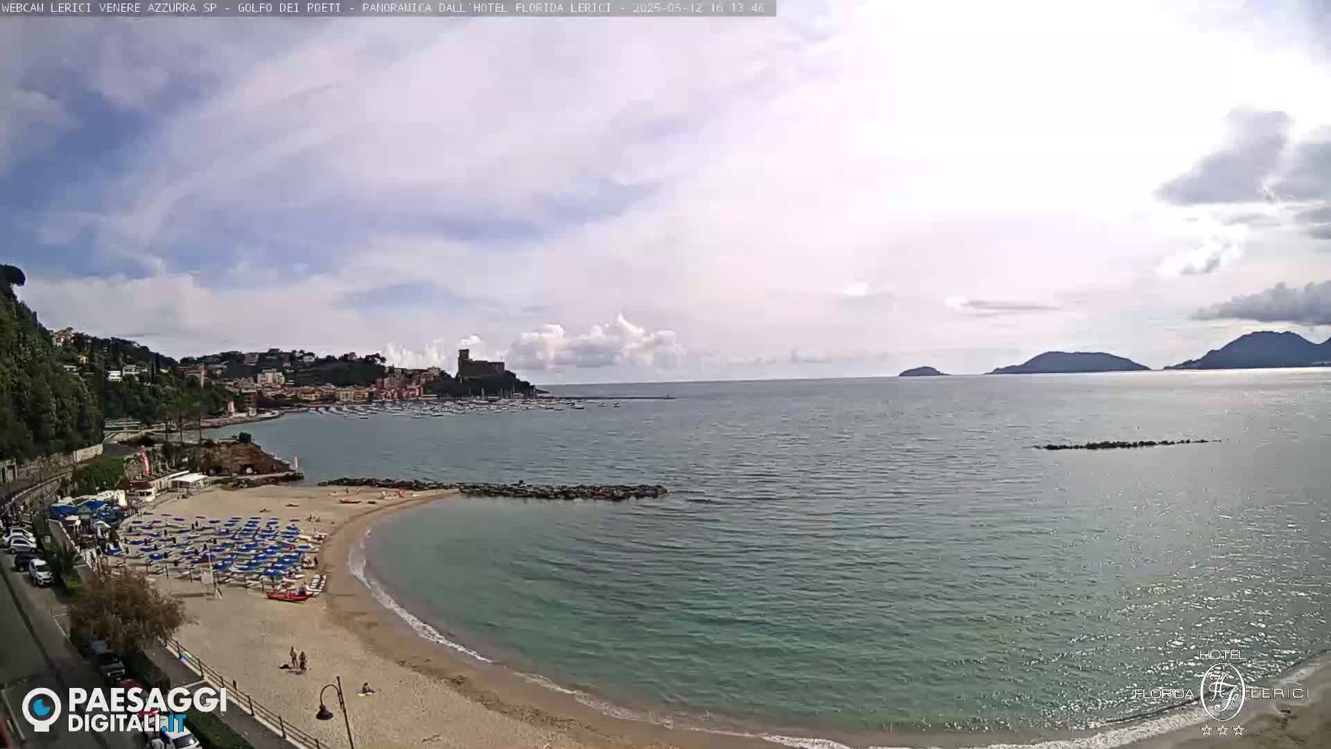 A partly cloudy day shows a sandy beach with umbrellas and chairs, a calm bay with boats, and a town nestled against hills, all overlooking a larger body of water with distant islands.