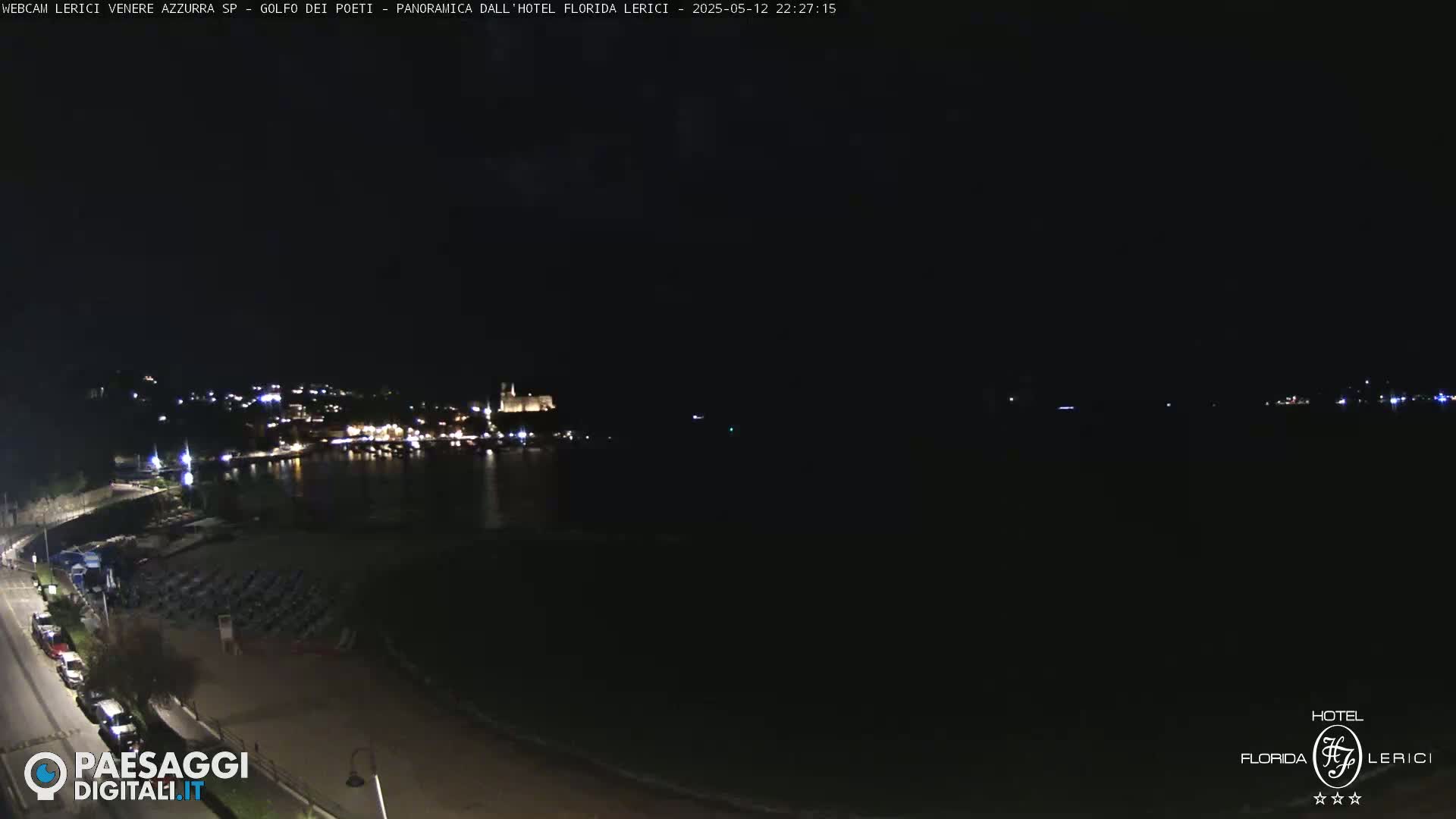 A nighttime view of a coastline with a town's lights across the water and a beach with rows of chairs in the foreground.