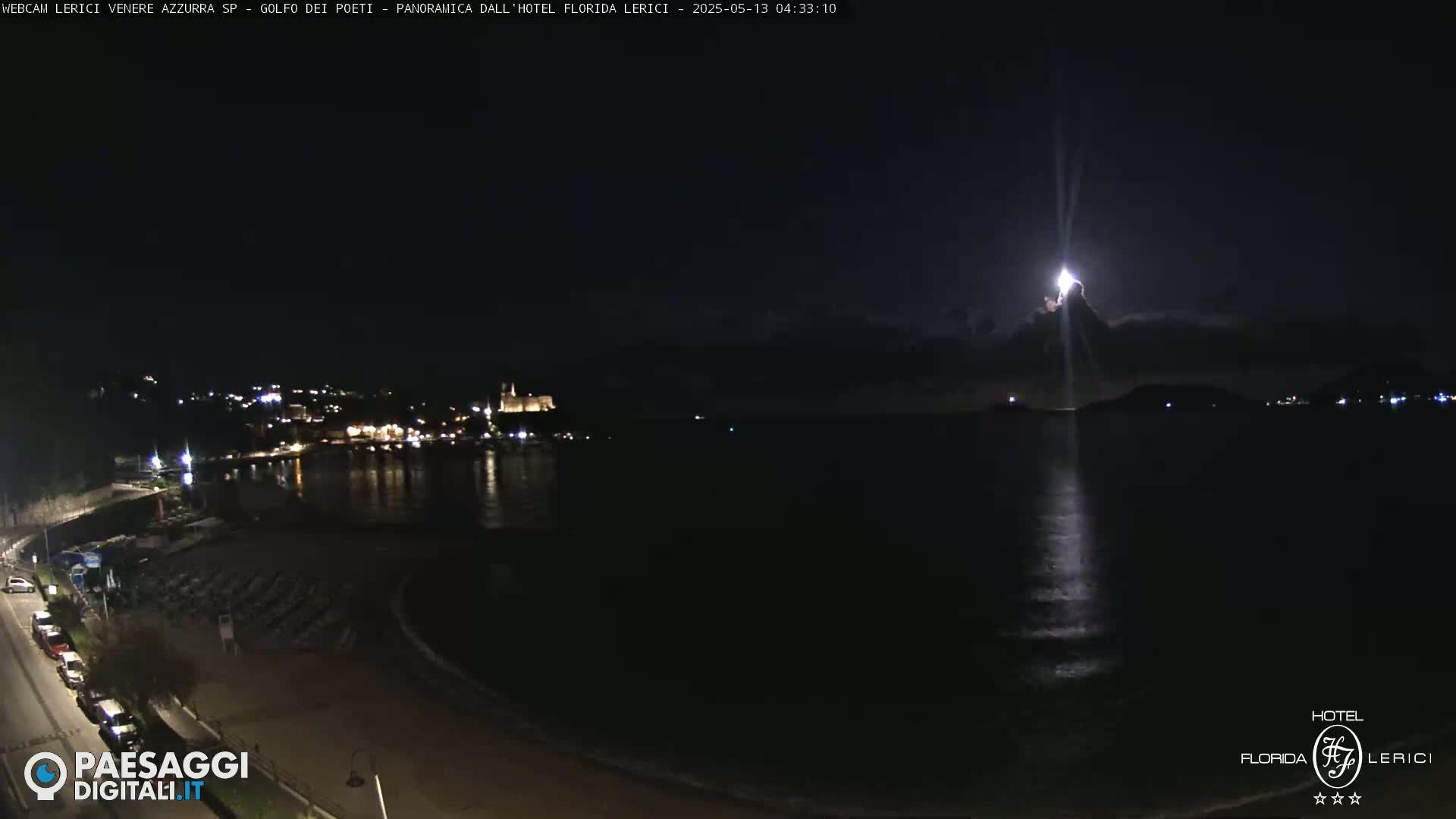 A nighttime coastal scene shows a moonlit body of water reflecting light, with a town's lights visible in the distance across the water.