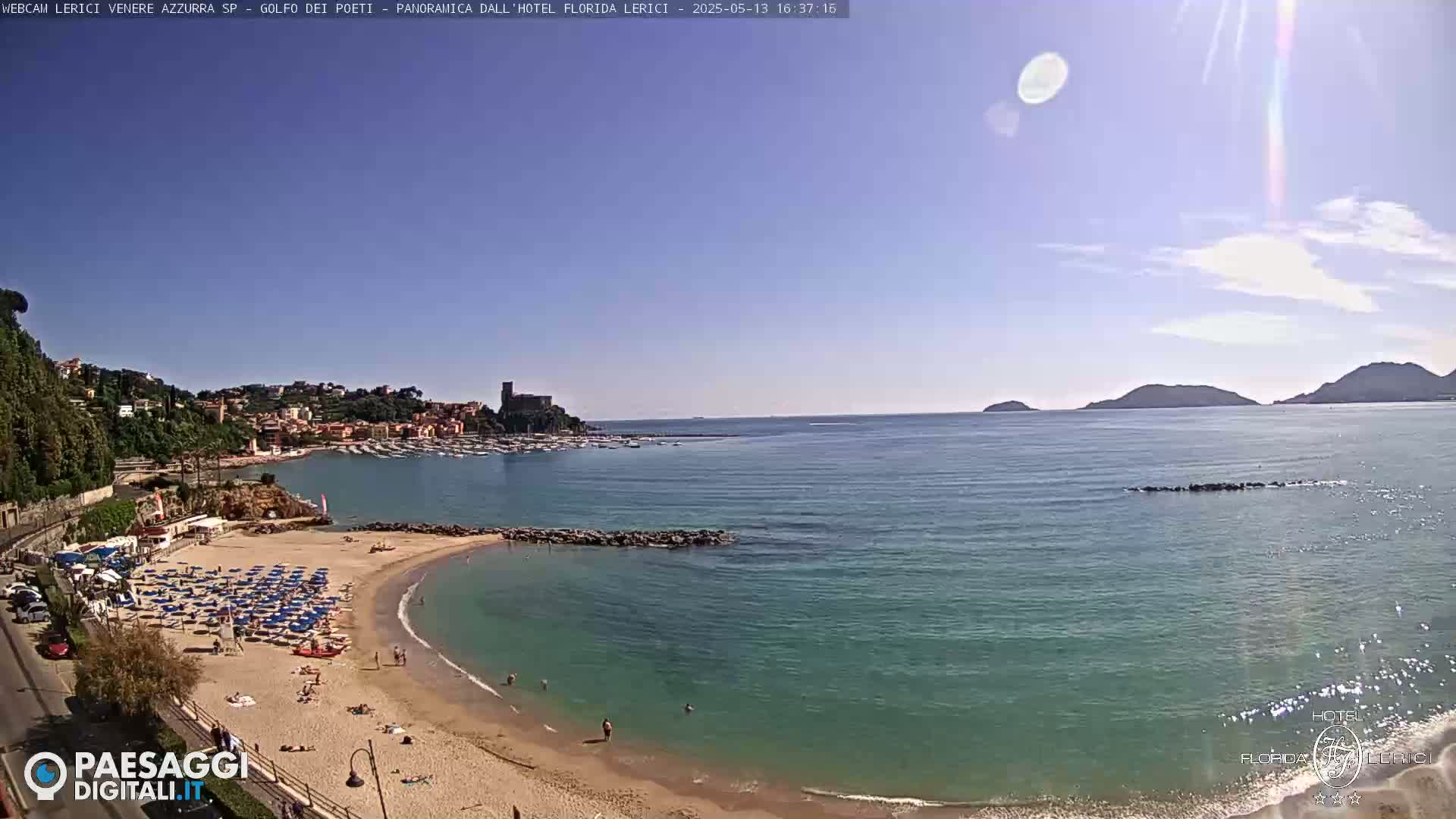 A sunny day at a sandy beach with calm, teal water, numerous beach umbrellas, and a town nestled on a hillside in the background.