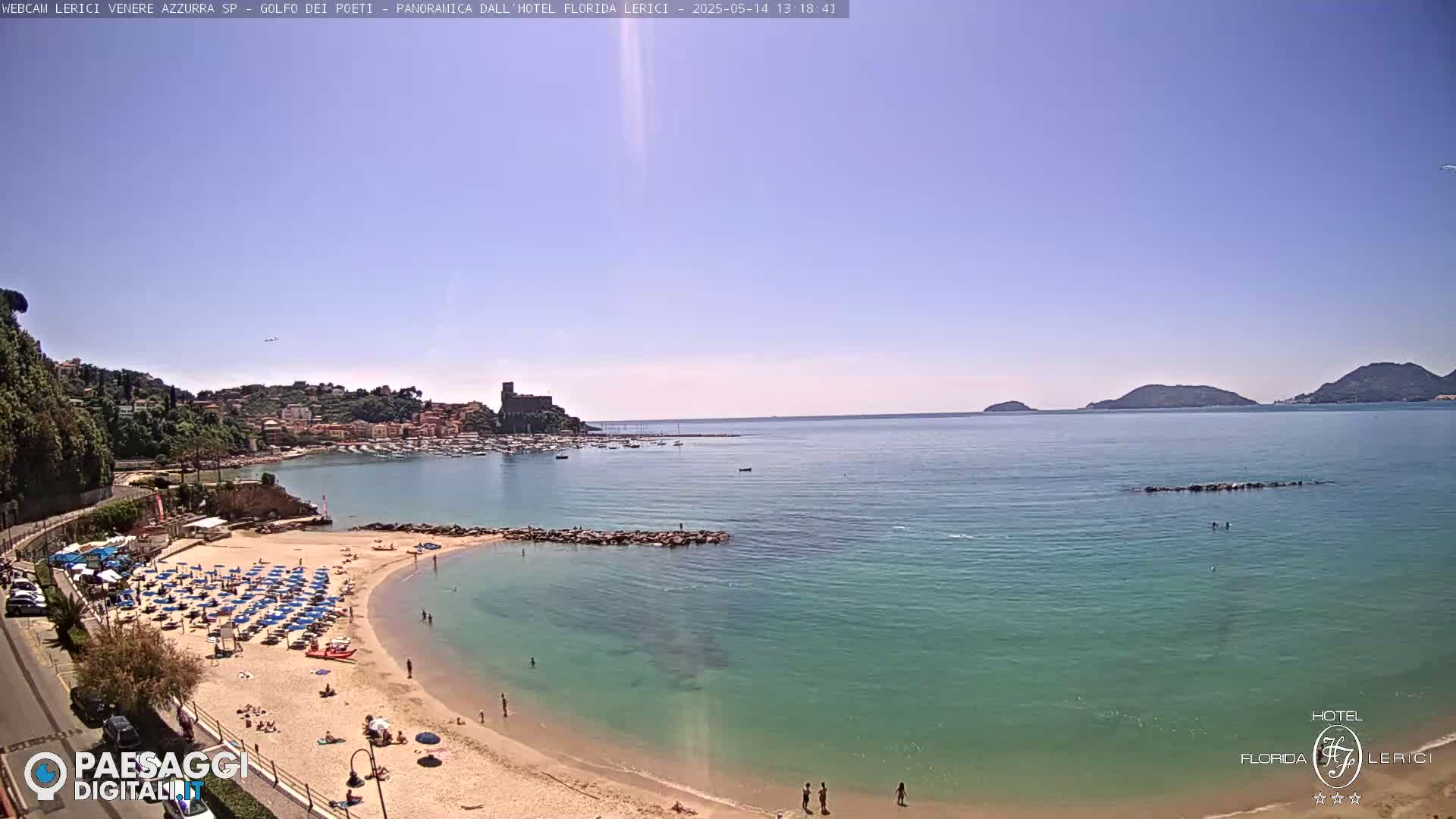 A sunny day at a beach with clear, teal water, showing a sandy shore lined with umbrellas and people, a small town nestled on a hillside across the bay, and distant islands under a clear sky.